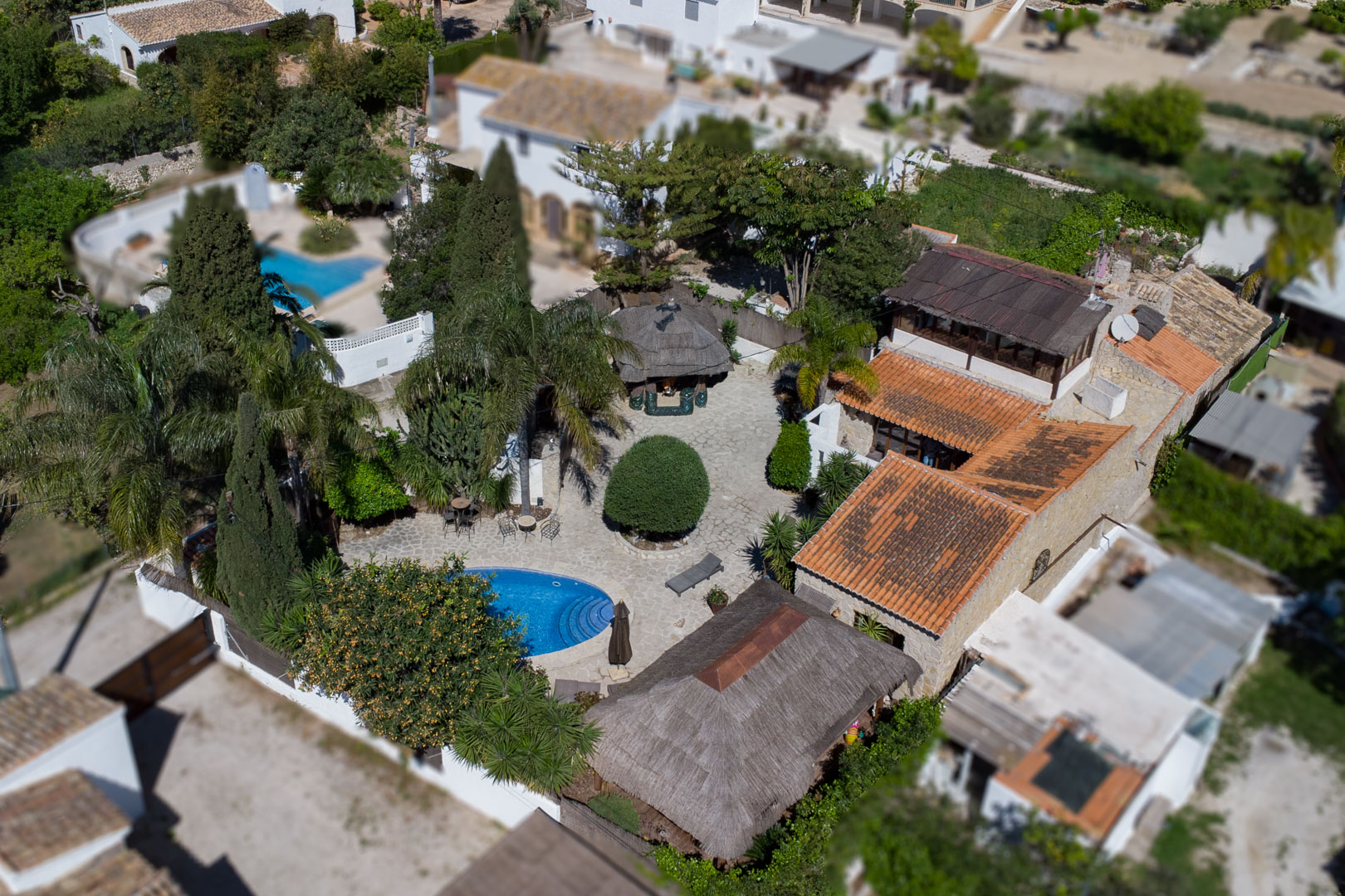 Vista aérea de la propiedad con piscina ovalada, patio de piedra natural y pérgolas balinesas rodeadas de vegetación mediterránea y muros de piedra.