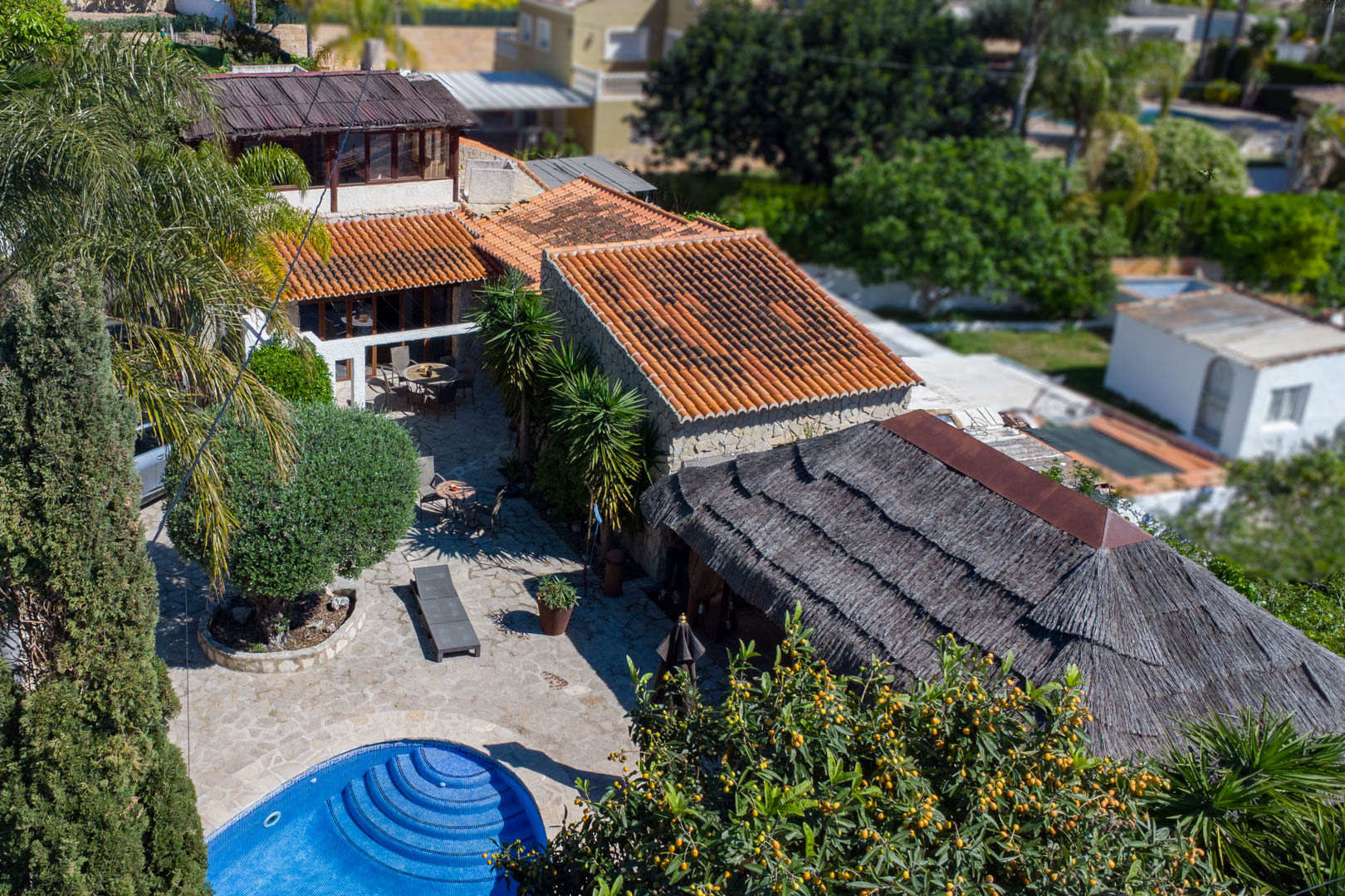 Vista aérea de la villa rústica con tejados de teja, piscina circular de mosaico azul, amplia terraza de piedra y una gran pérgola balinesa rodeada de vegetación.