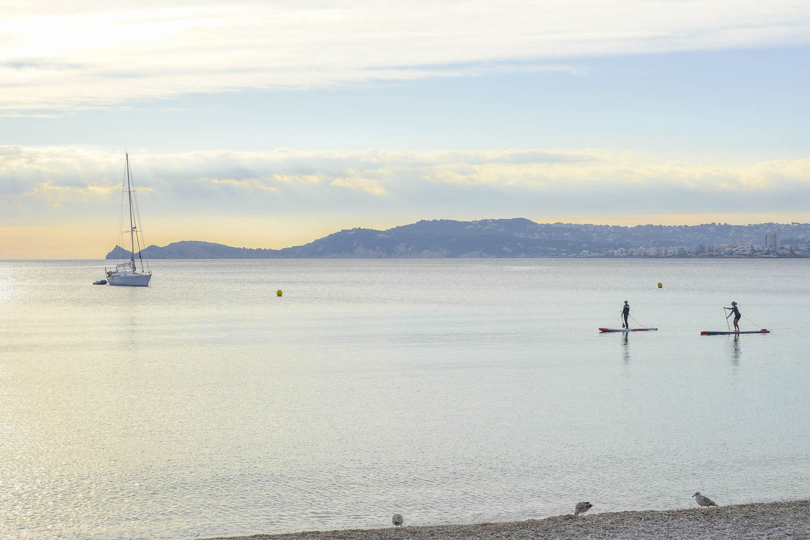 Vista panorámica de la bahía con aguas tranquilas, un velero fondeado y practicantes de paddle surf frente a la costa de Jávea.