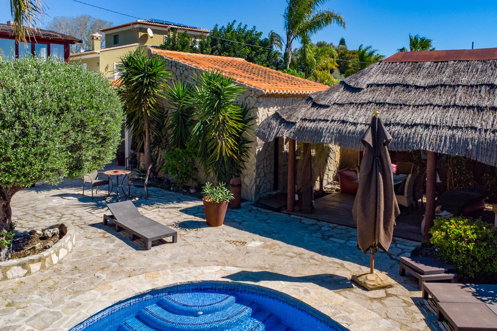 Zona exterior con patio de piedra natural, piscina y una pérgola balinesa de brezo junto a una construcción de piedra rústica y vegetación mediterránea.