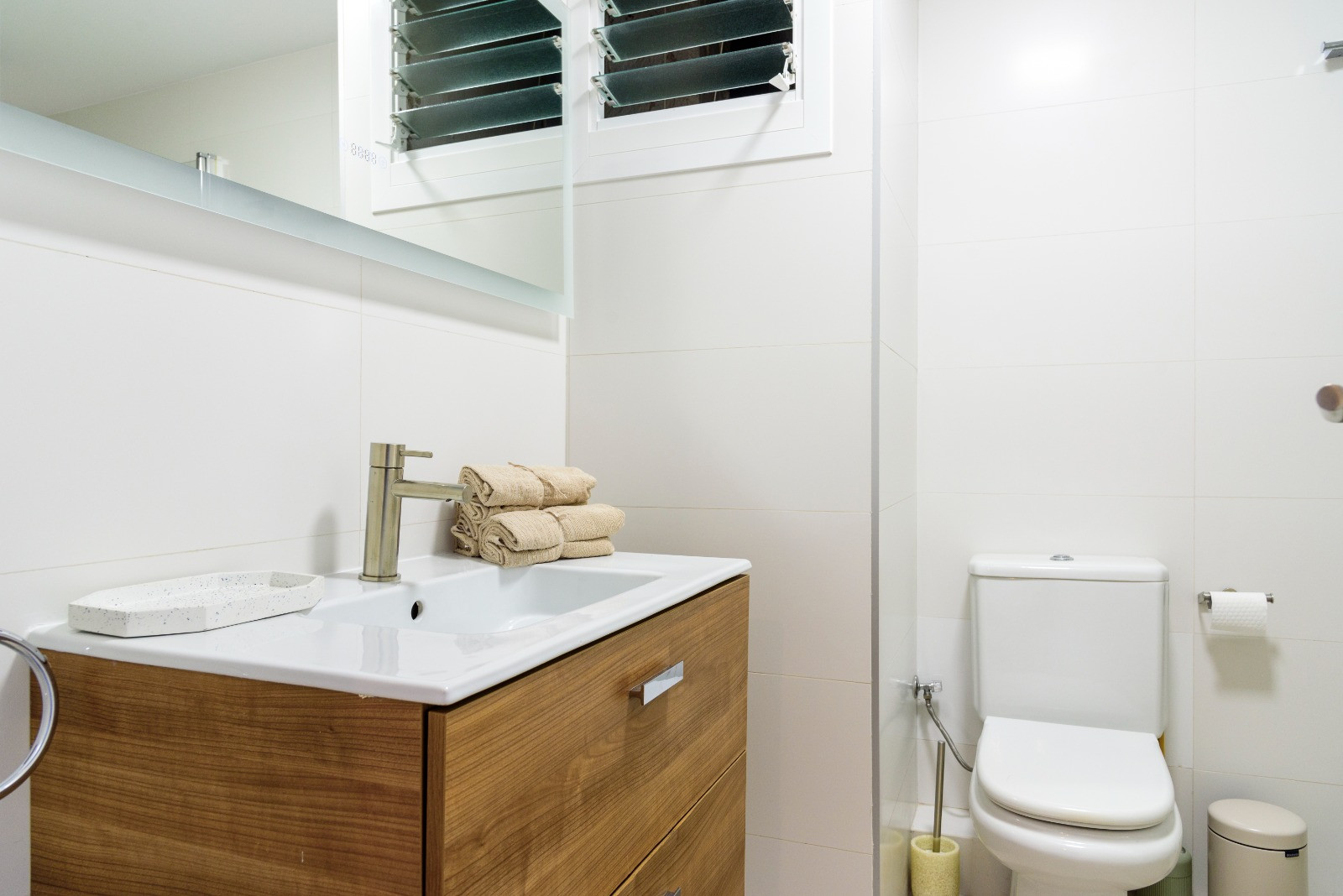 Baño moderno con mueble de madera, lavabo integrado y paredes revestidas en blanco. Incluye sanitarios actuales y una ventana de lamas para ventilación natural.