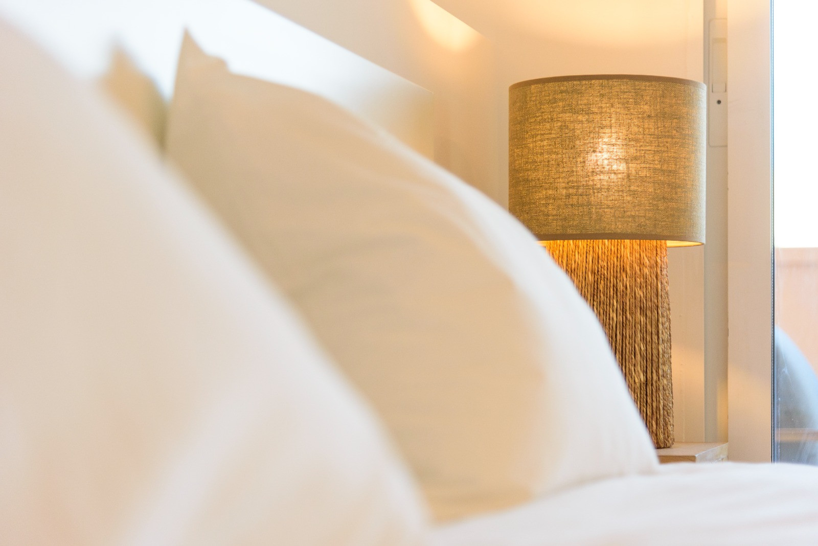 Detalle de dormitorio con lámpara de sobremesa de fibras naturales y pantalla de lino, aportando calidez junto a la cama con textiles blancos y luz natural.