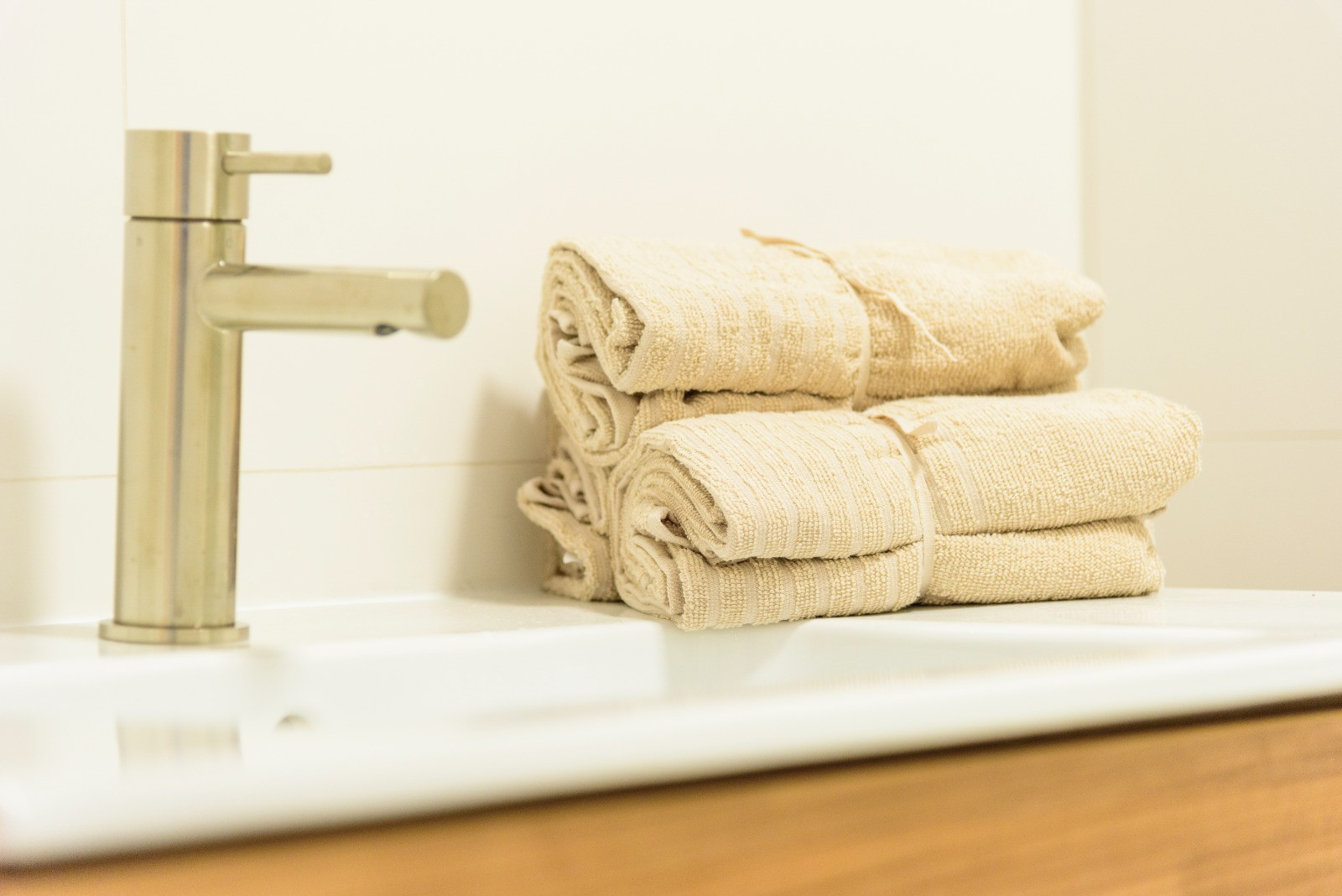 Detalle del baño con grifería de acero cepillado, lavabo blanco y toallas de algodón en tonos neutros sobre un mueble de madera contemporáneo.