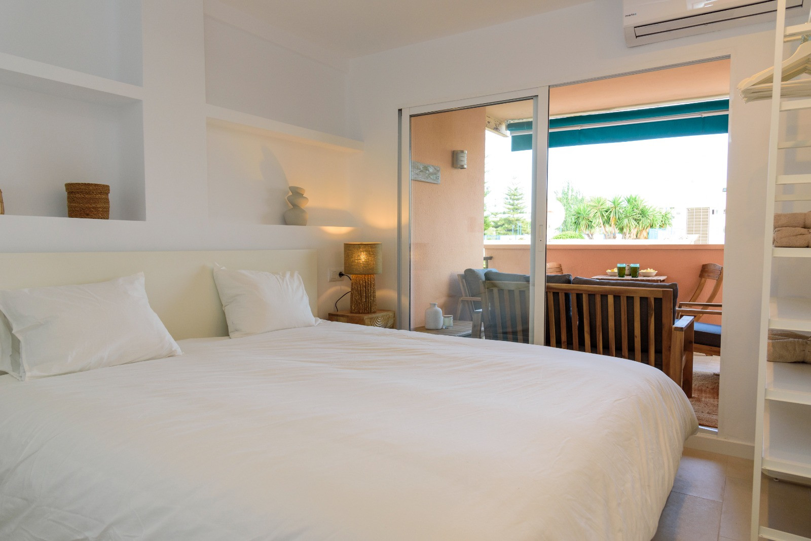 Dormitorio con estanterías integradas y acceso directo a una terraza privada equipada con mobiliario de madera, creando un ambiente fluido y luminoso.