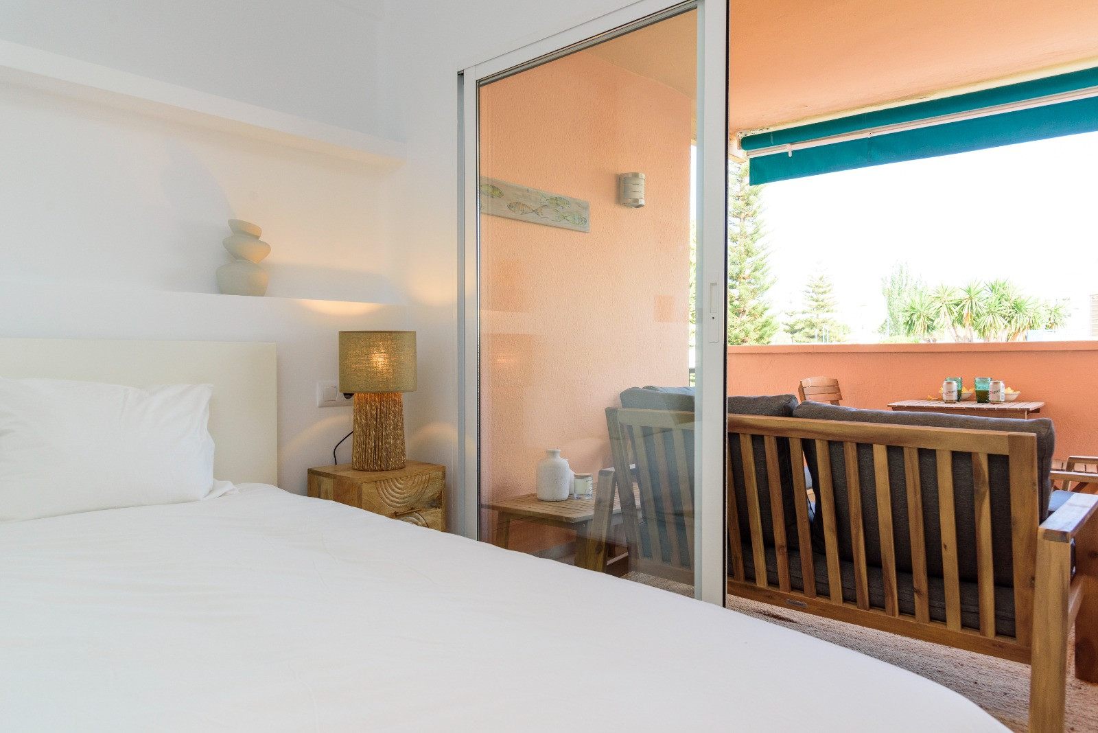 Dormitorio luminoso con acceso directo a una terraza privada equipada con mobiliario de madera. Destaca la luz natural y el diseño minimalista con tonos neutros.
