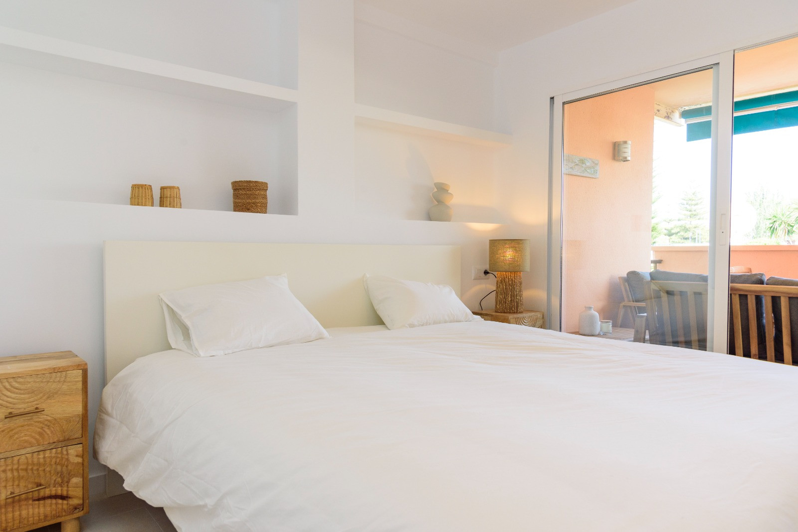 Dormitorio luminoso con estanterías integradas en la pared, mesitas de noche de madera y acceso directo a la terraza privada a través de grandes ventanales correderos.