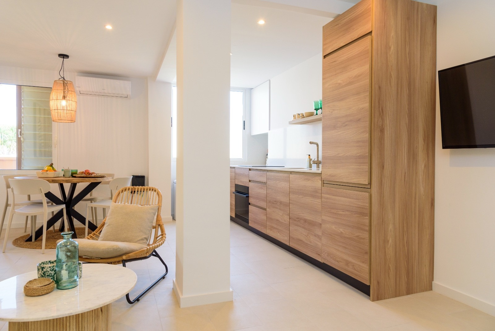 Zona de día diáfana con cocina integrada de madera, comedor con lámpara de fibras naturales y suelos cerámicos claros bajo iluminación empotrada.
