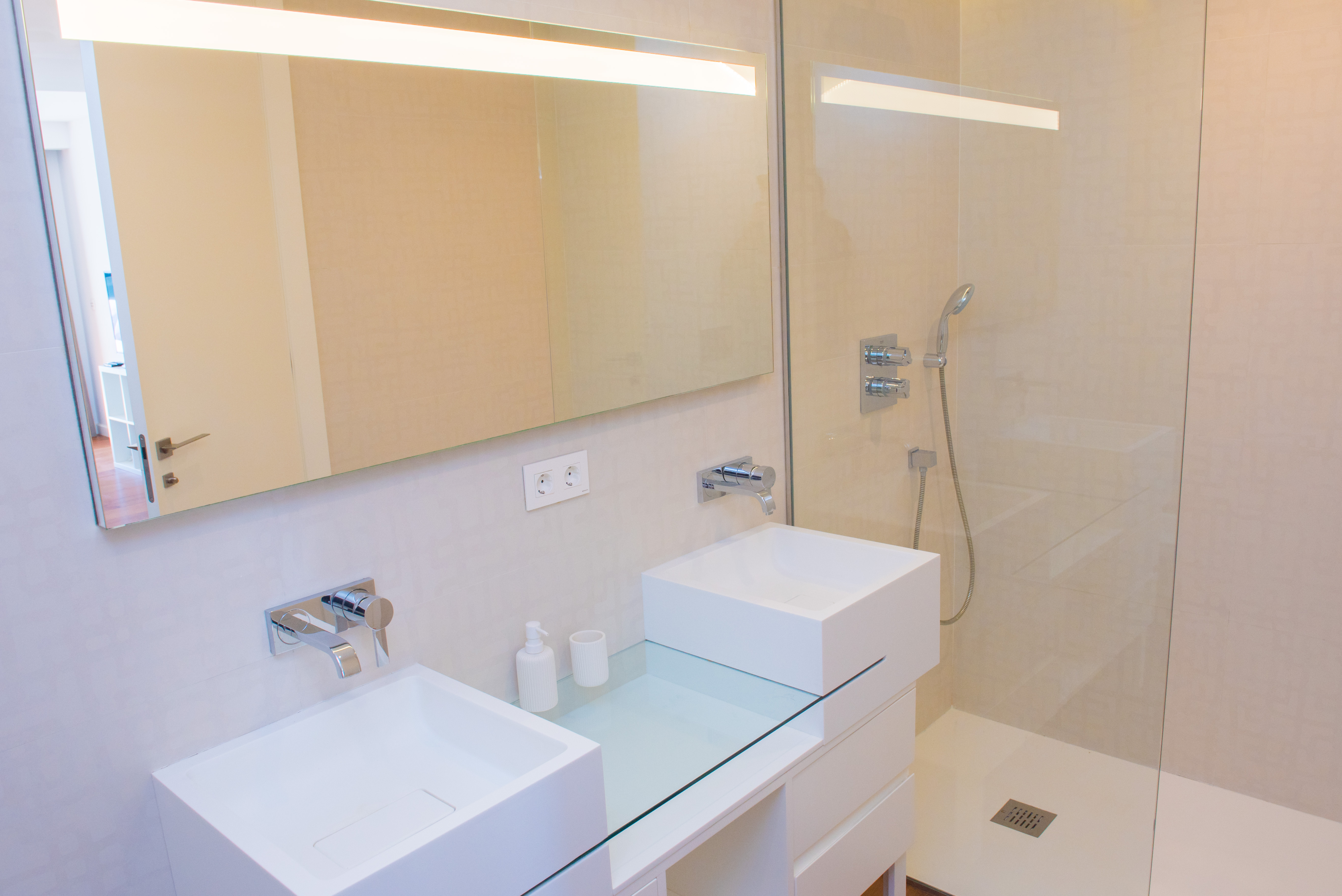 Baño contemporáneo con lavabo doble de diseño cuadrado, grifería cromada empotrada y ducha a ras de suelo con mampara de cristal y espejo retroiluminado.