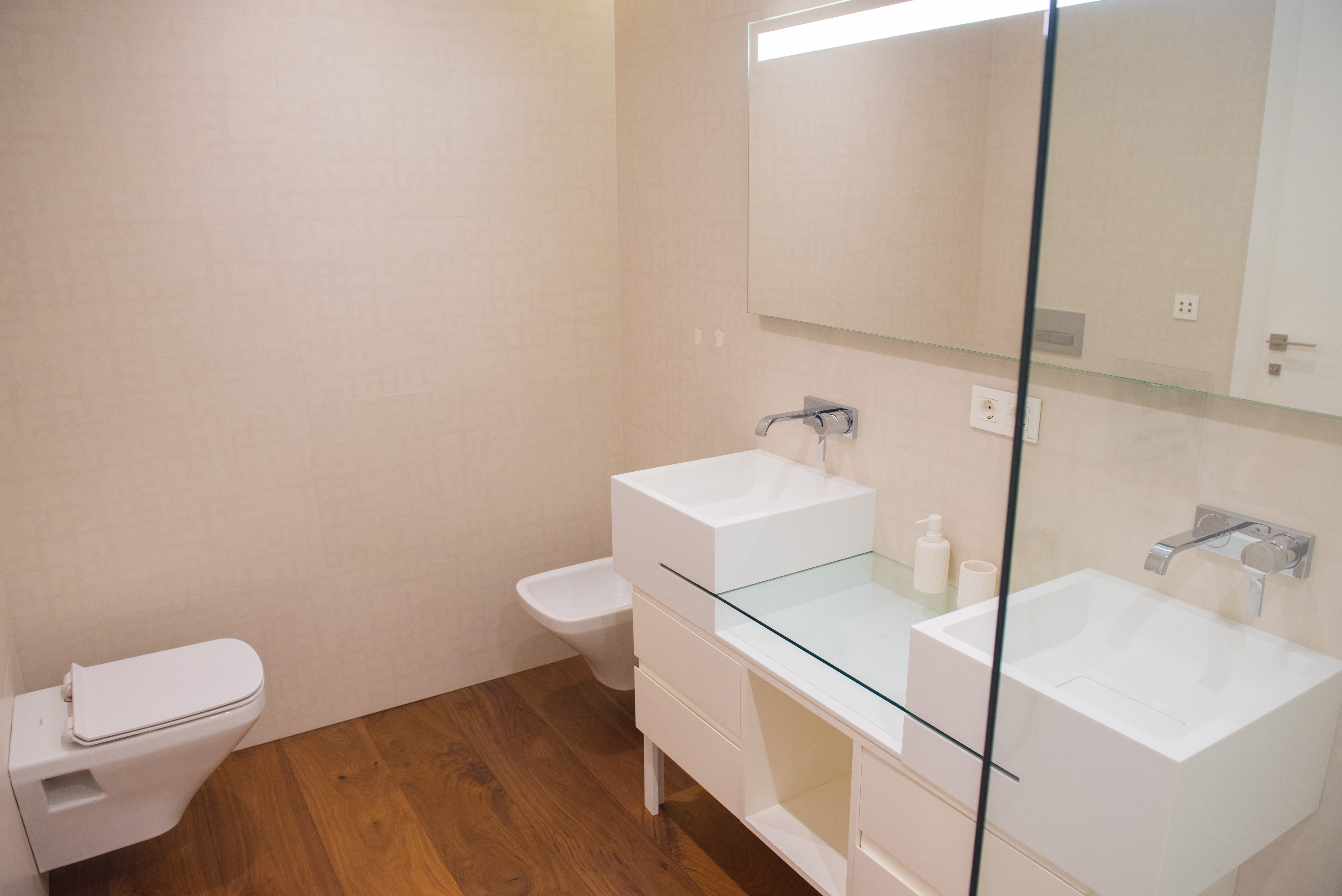 Baño contemporáneo con lavabo doble de seno cuadrado, encimera de cristal y grifería empotrada. Dispone de sanitarios suspendidos y suelo de madera.