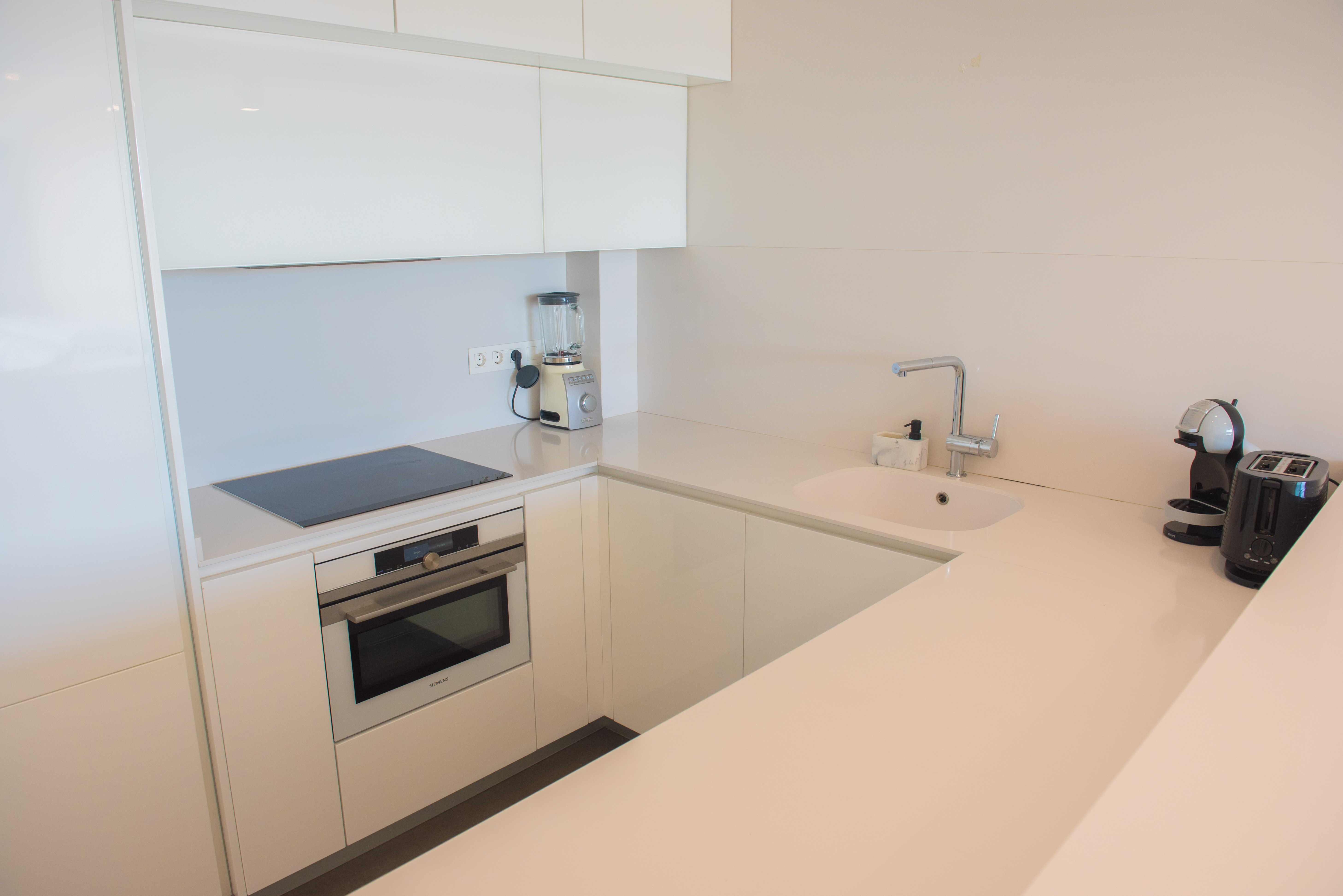 Cocina minimalista en blanco brillante con encimera de cuarzo, equipada con horno Siemens, placa de inducción y electrodomésticos de alta gama integrados.