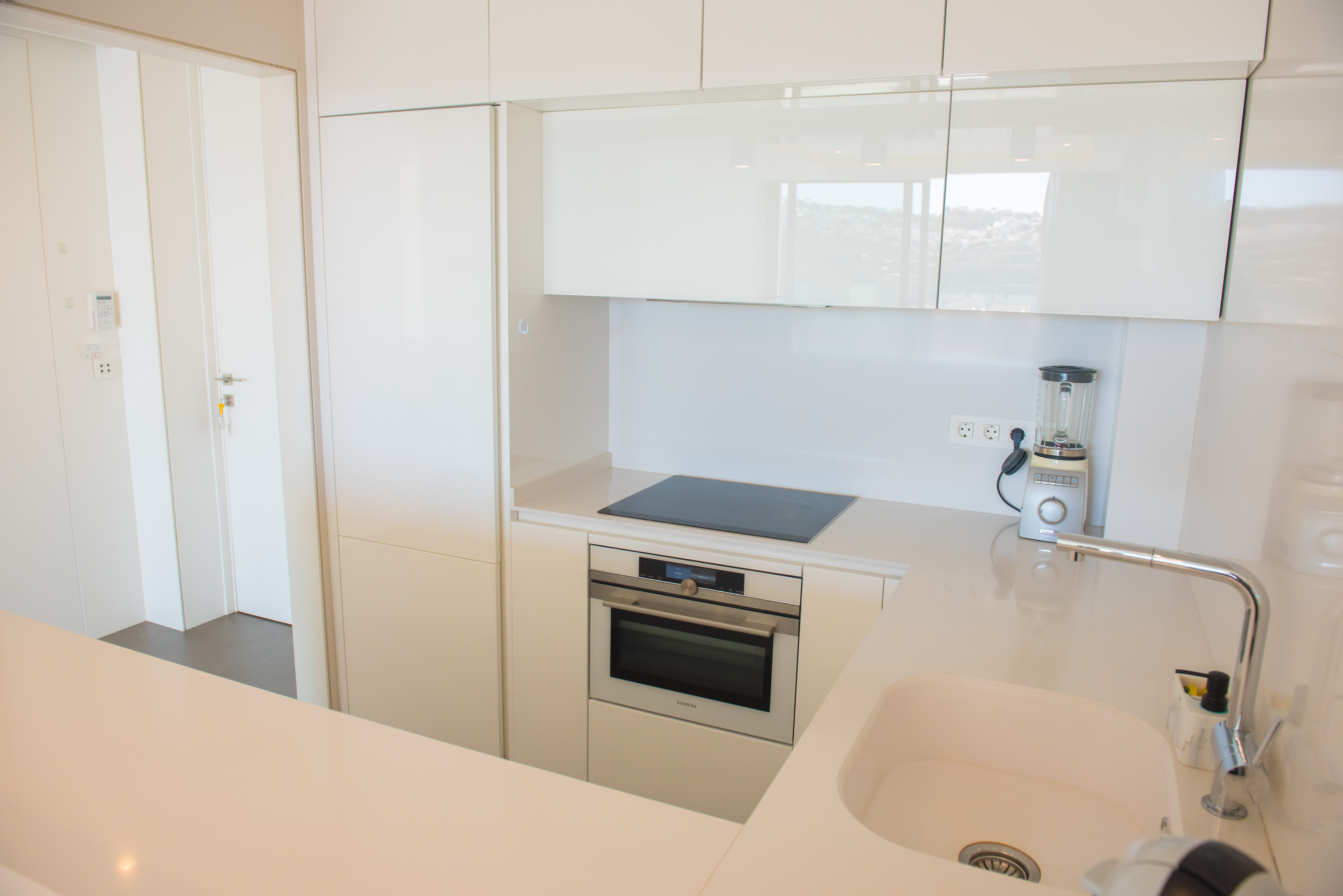 Cocina moderna con armarios en blanco brillante, placa de inducción, horno Siemens integrado y encimera de cuarzo blanco con fregadero bajo encimera.