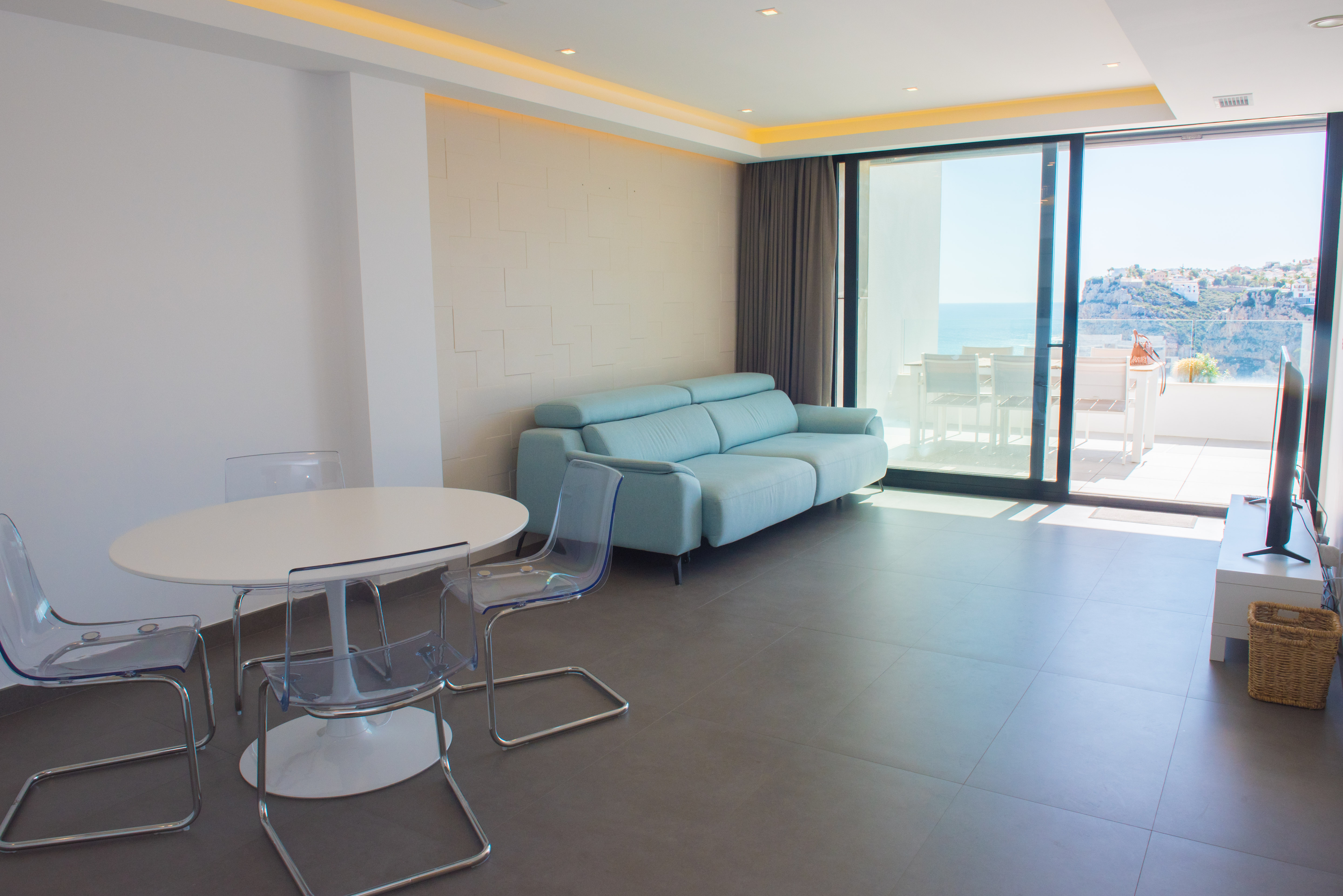Salón-comedor con sofá azul pastel, mesa ovalada y sillas acrílicas. Destaca la pared texturizada, iluminación indirecta y acceso directo a la terraza con vistas al mar.
