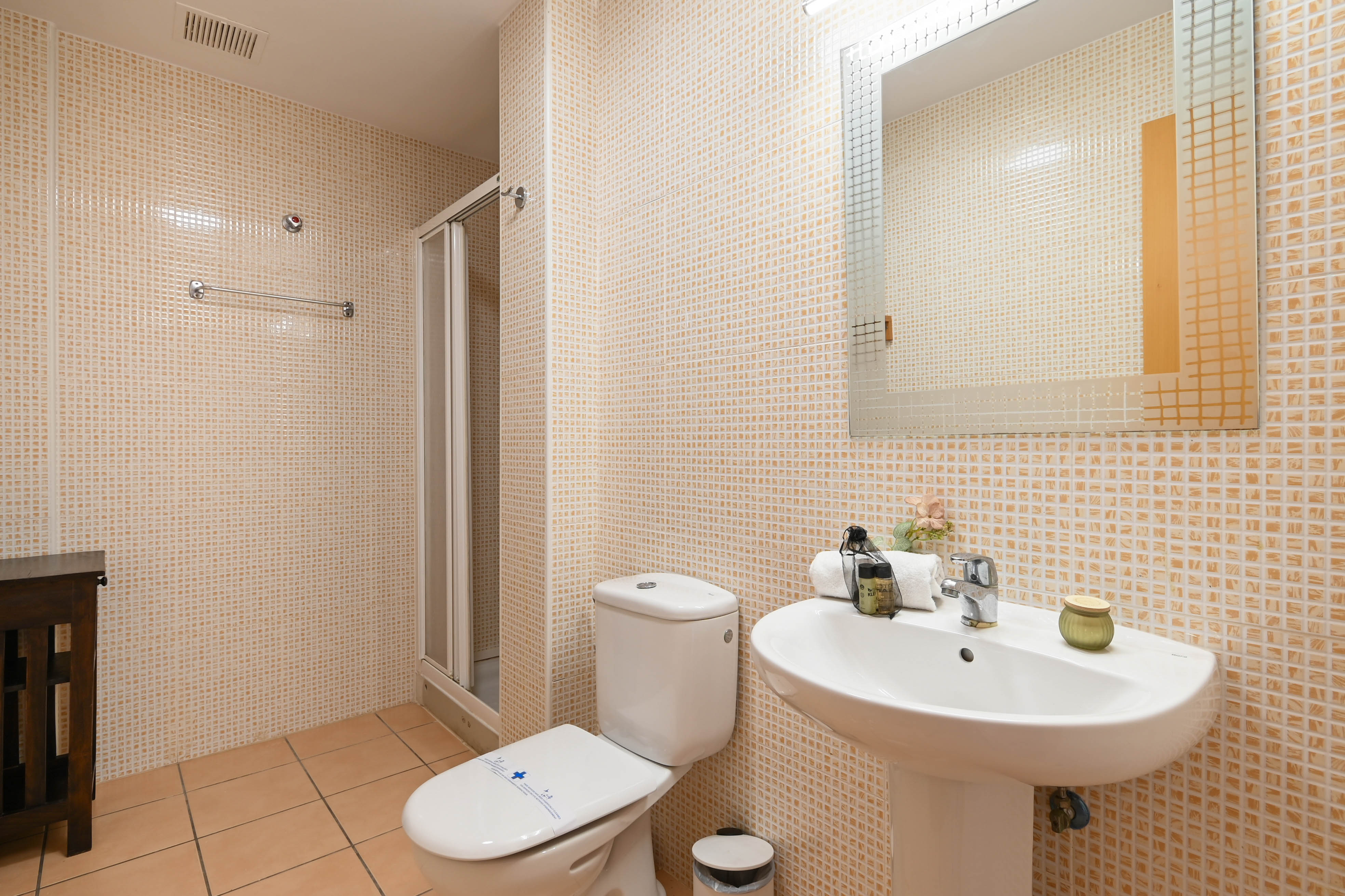 Baño funcional con paredes revestidas en gresite beige, lavabo de pedestal blanco y una ducha con mampara plegable de cristal.