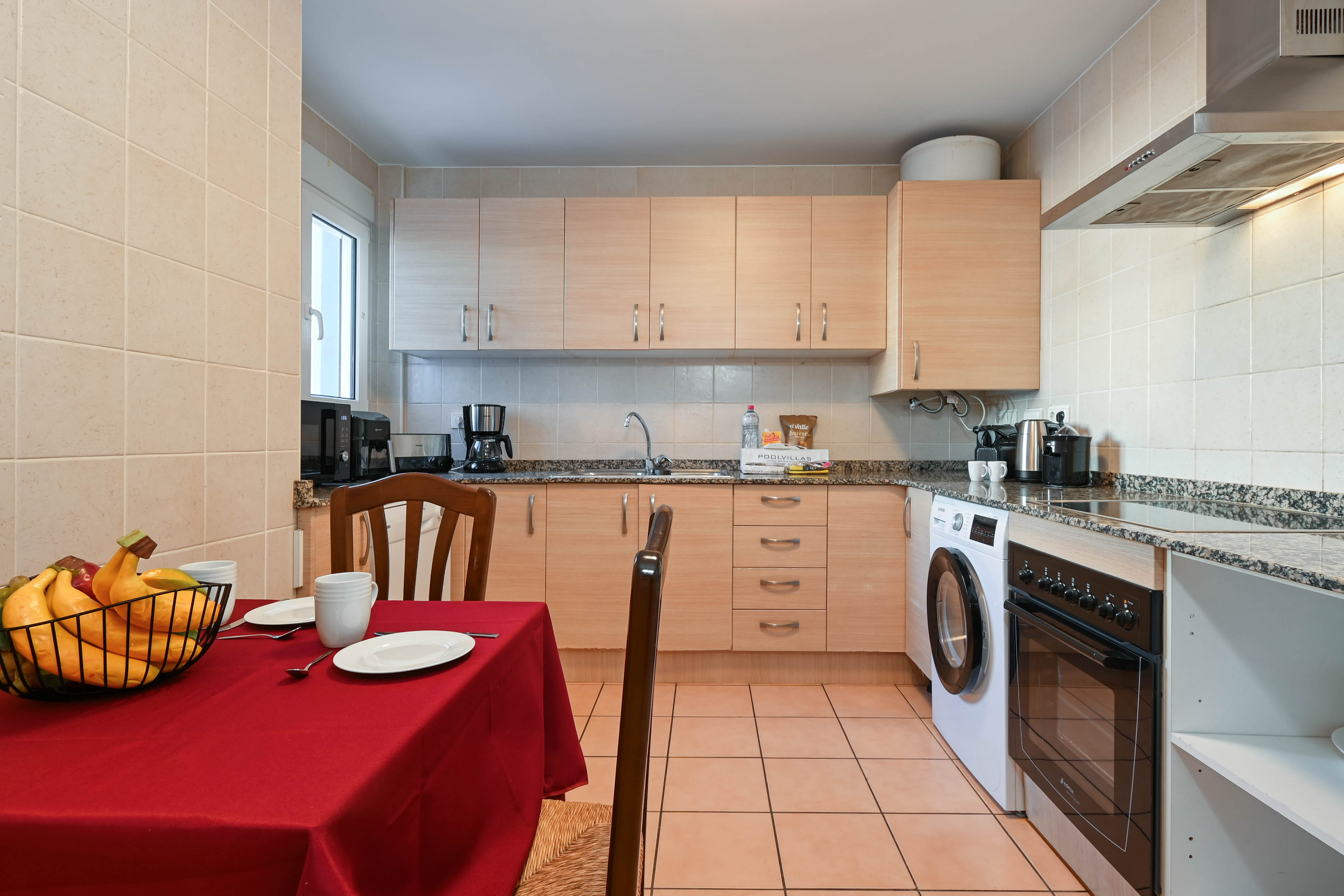 Cocina equipada con armarios de madera clara, encimeras de granito y electrodomésticos integrados, junto a una zona de comedor con mantel rojo.