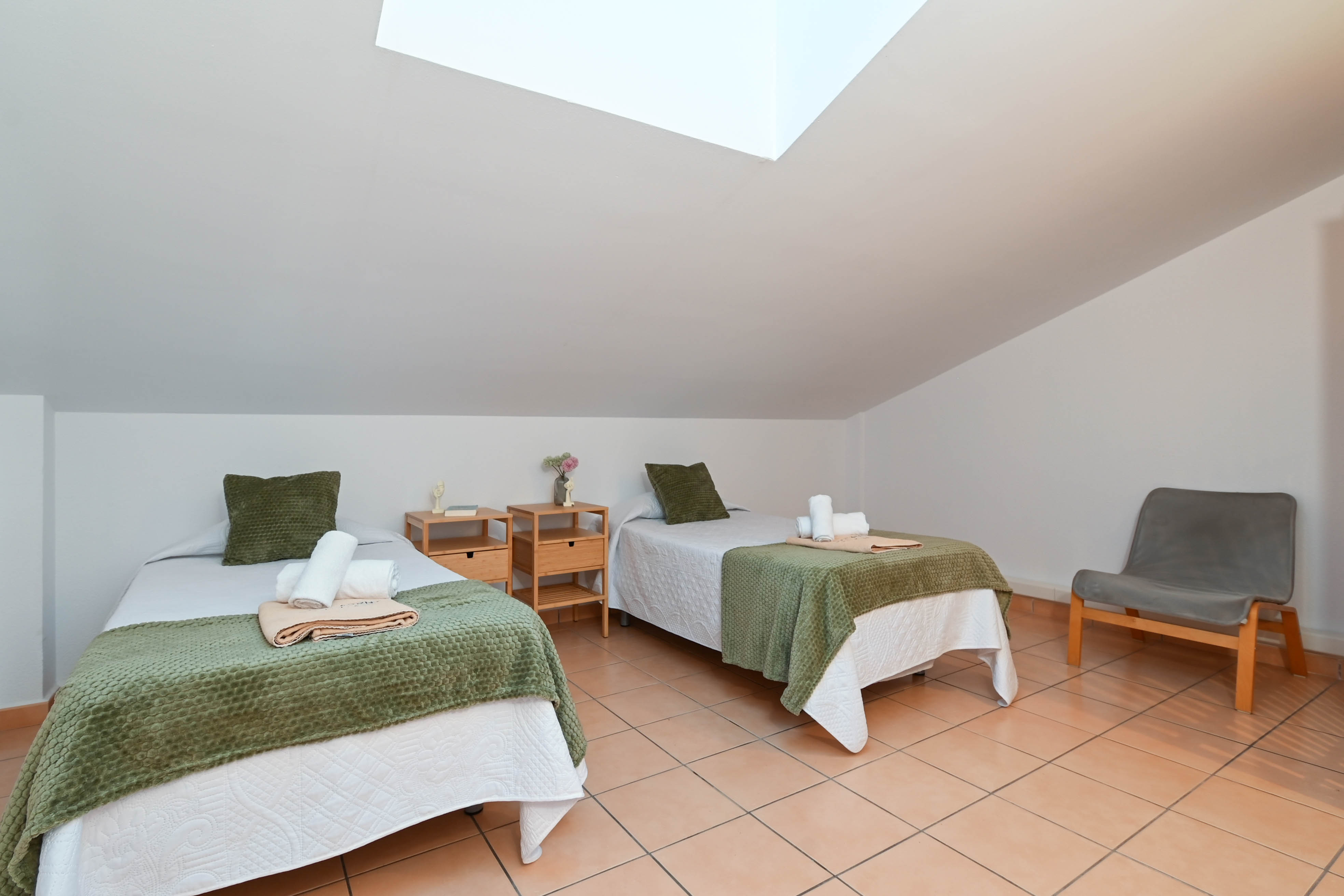 Dormitorio abuhardillado con dos camas individuales, suelos de terracota y una claraboya que proporciona luz natural directa al espacio.