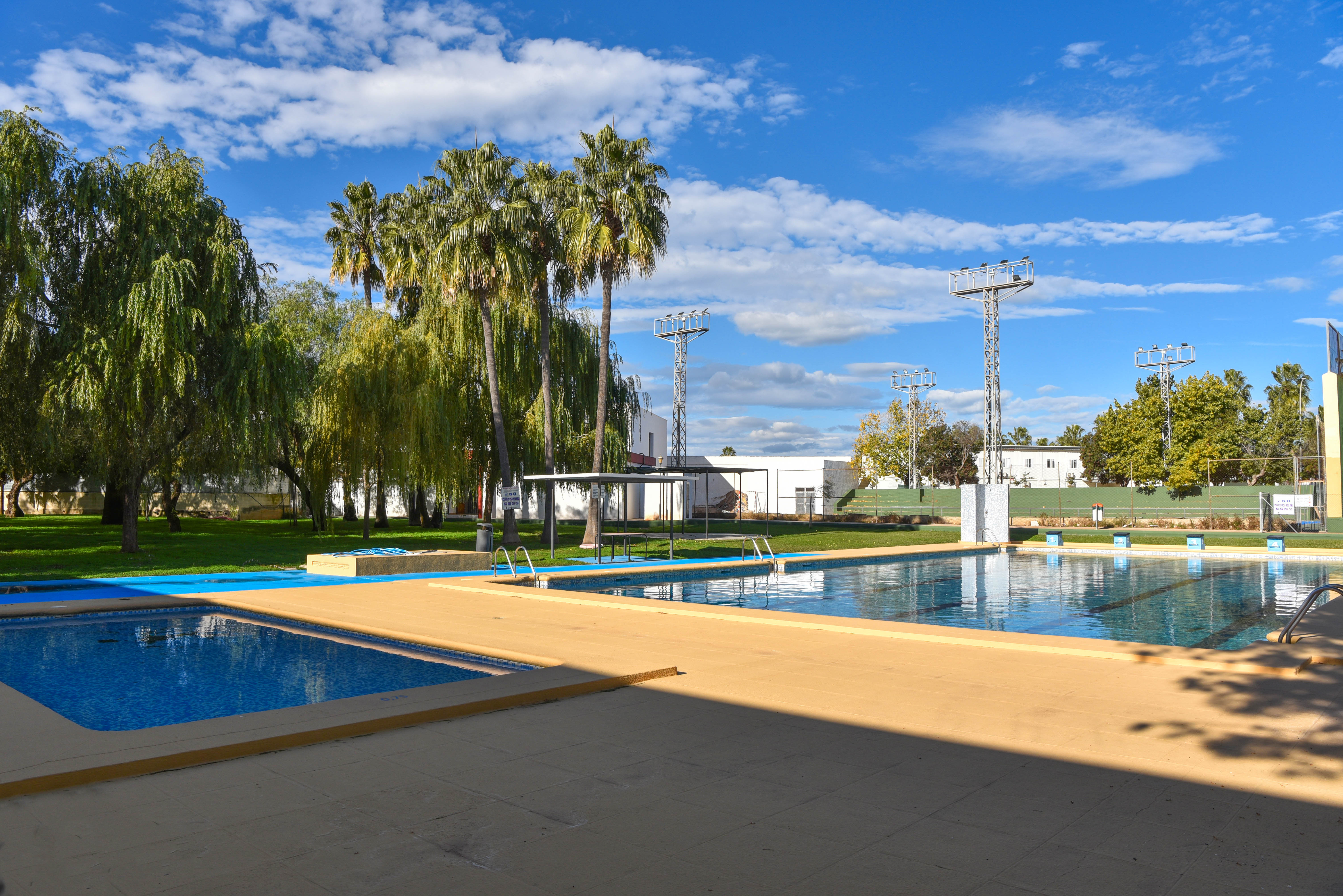 Piscina municipal con zona infantil, amplia terraza solárium y áreas verdes con palmeras, situada bajo torres de iluminación deportiva.