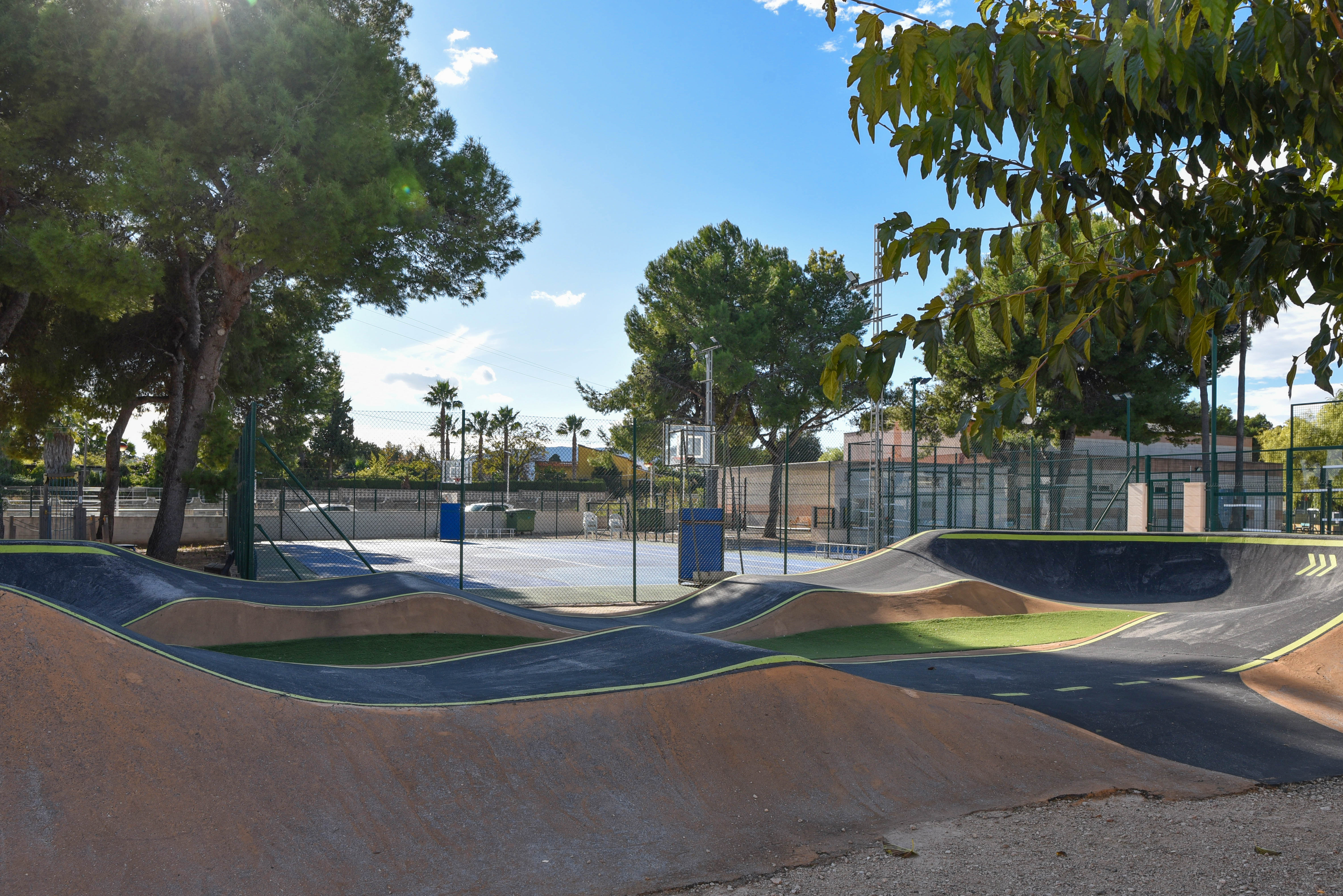 Pista de pump track con ondulaciones de asfalto junto a una cancha polideportiva vallada, rodeada de pinos maduros en una zona recreativa municipal.