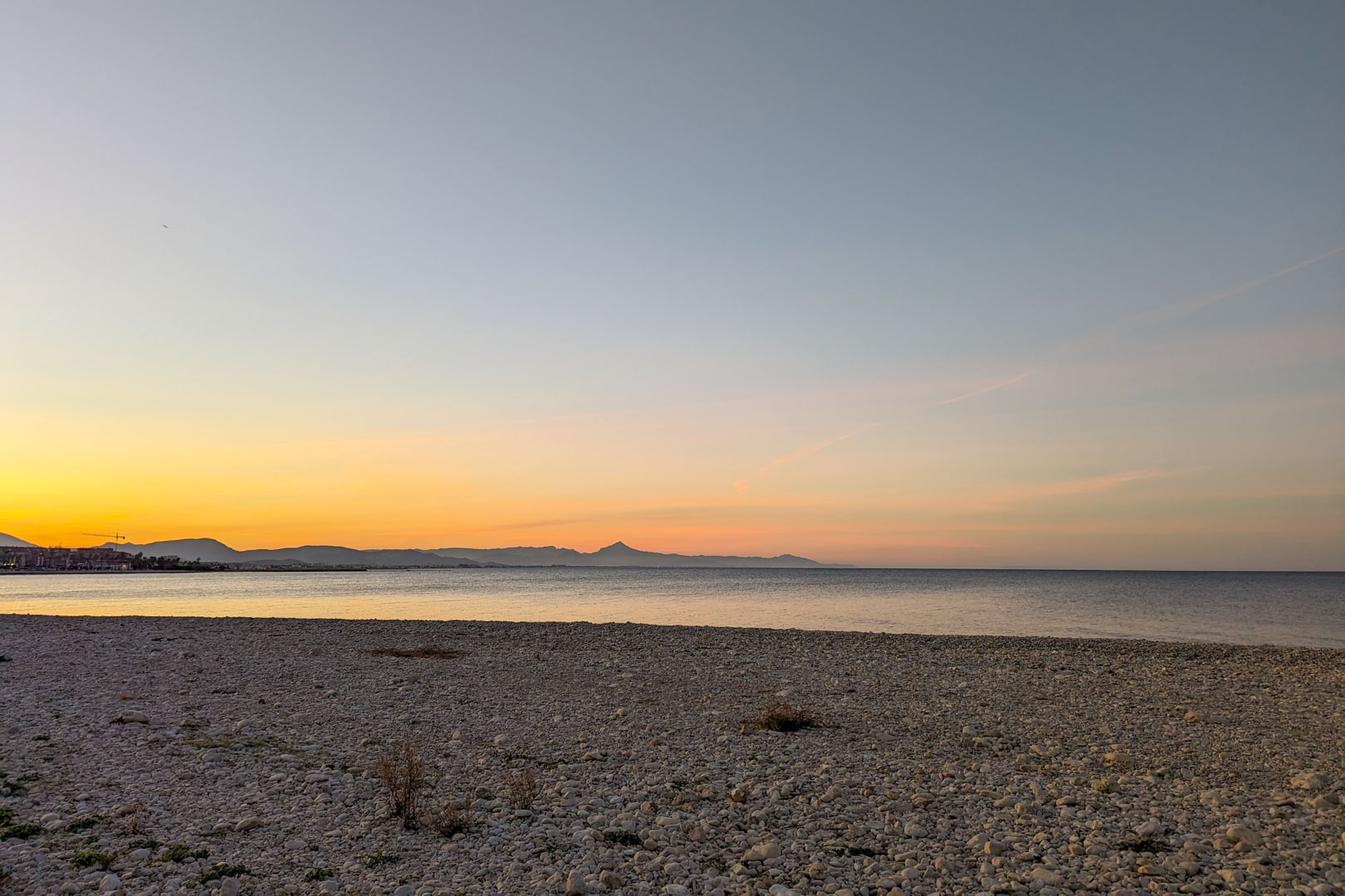 Vista panorámica de la playa de cantos rodados cercana a la vivienda durante el atardecer, con siluetas de montañas en el horizonte y aguas tranquilas.