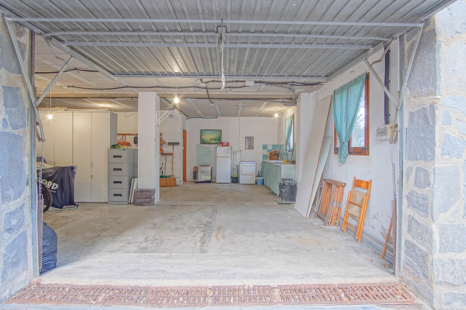 Amplio semisótano con capacidad para varios vehículos, zona de taller y cocina integrada. Presenta suelos de hormigón, pilares de piedra y acceso directo mediante portón metálico.