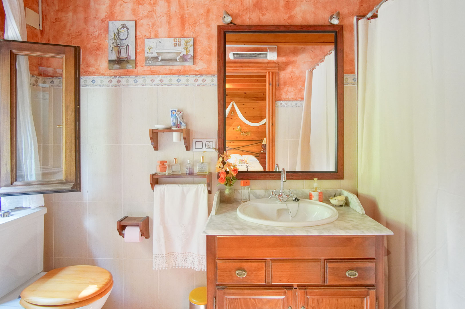 Baño de estilo rústico con mueble de lavabo en madera, encimera de mármol y paredes en tonos terracota, con un espejo que refleja el dormitorio revestido en madera de pino.