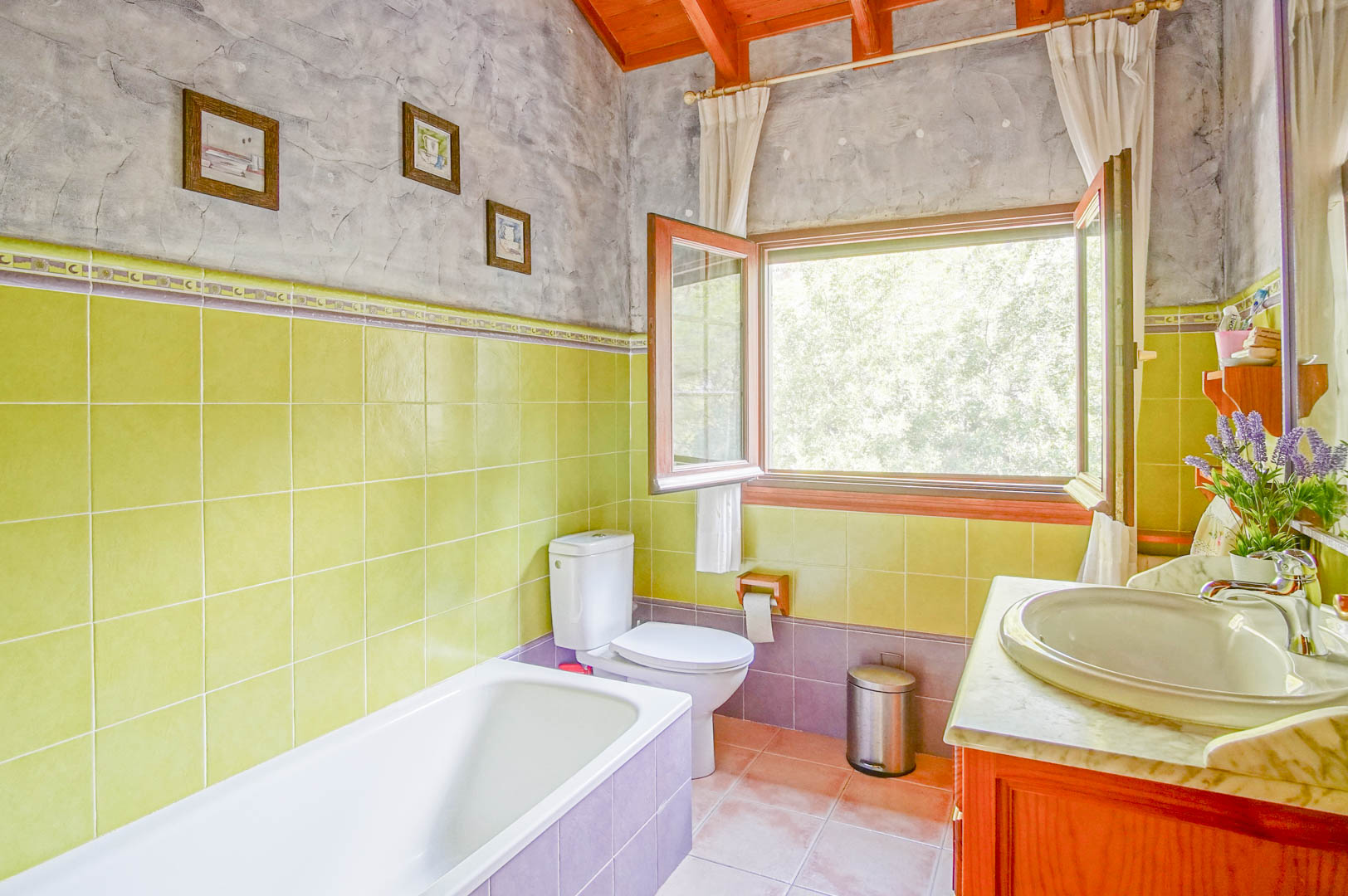Cuarto de baño con techos de vigas de madera, paredes de azulejos verdes y lavabo con encimera de mármol. Una gran ventana proporciona luz natural y vistas al entorno verde.