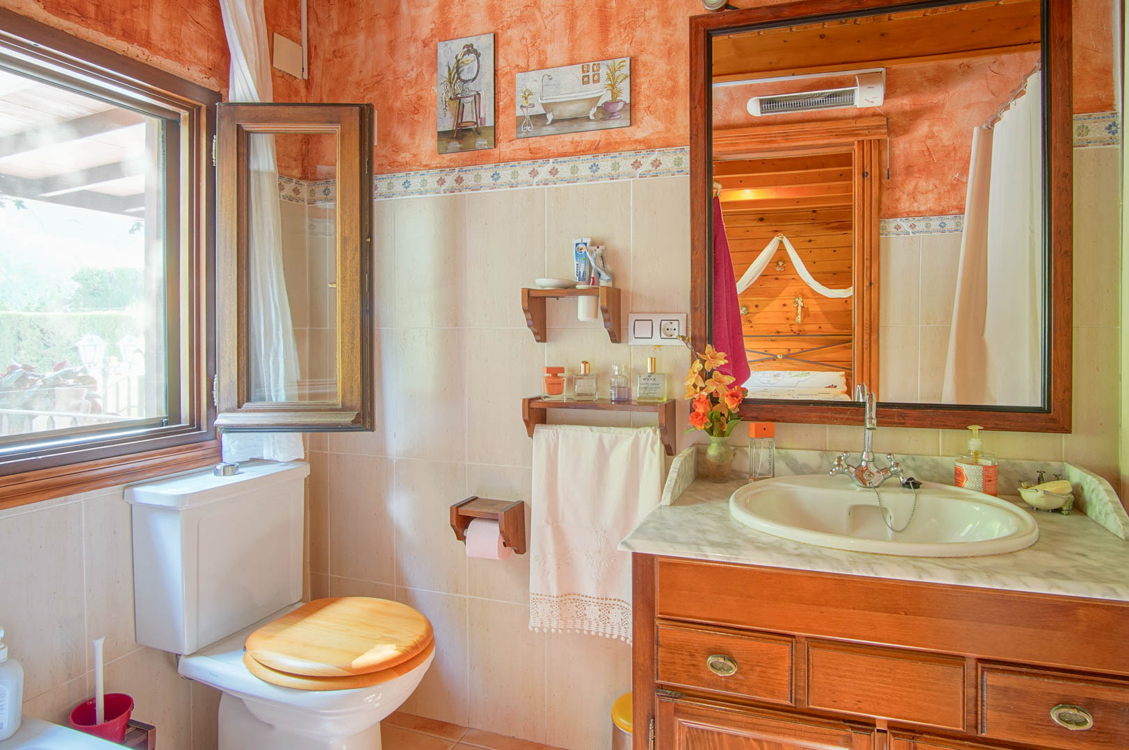 Cuarto de baño rústico con mueble de madera, encimera de mármol y ventana al exterior. El espejo refleja el dormitorio contiguo revestido en madera de pino.