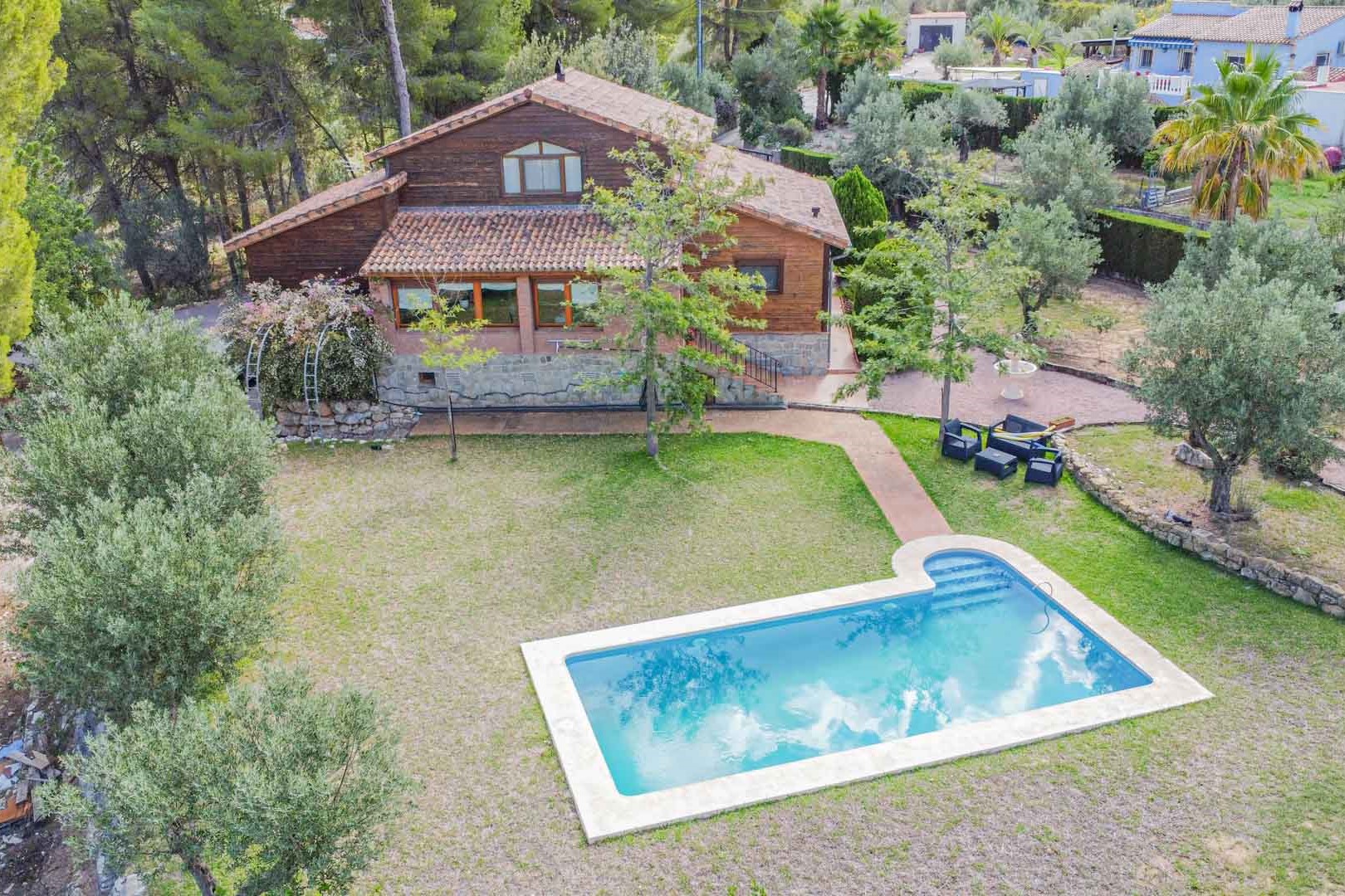 Vista aérea de la villa con fachada de madera y piedra, destacando la piscina privada rodeada de césped y jardines mediterráneos con olivos.