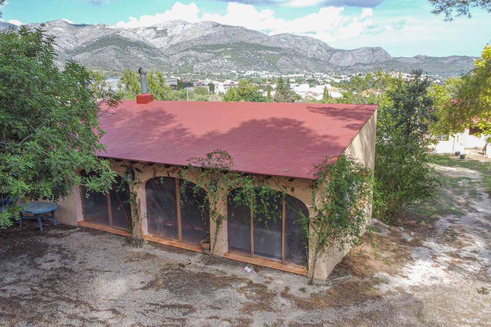 Vista exterior de la edificación anexa con arcos acristalados y chimenea, rodeada de vegetación mediterránea y con vistas panorámicas a las montañas de Orba.