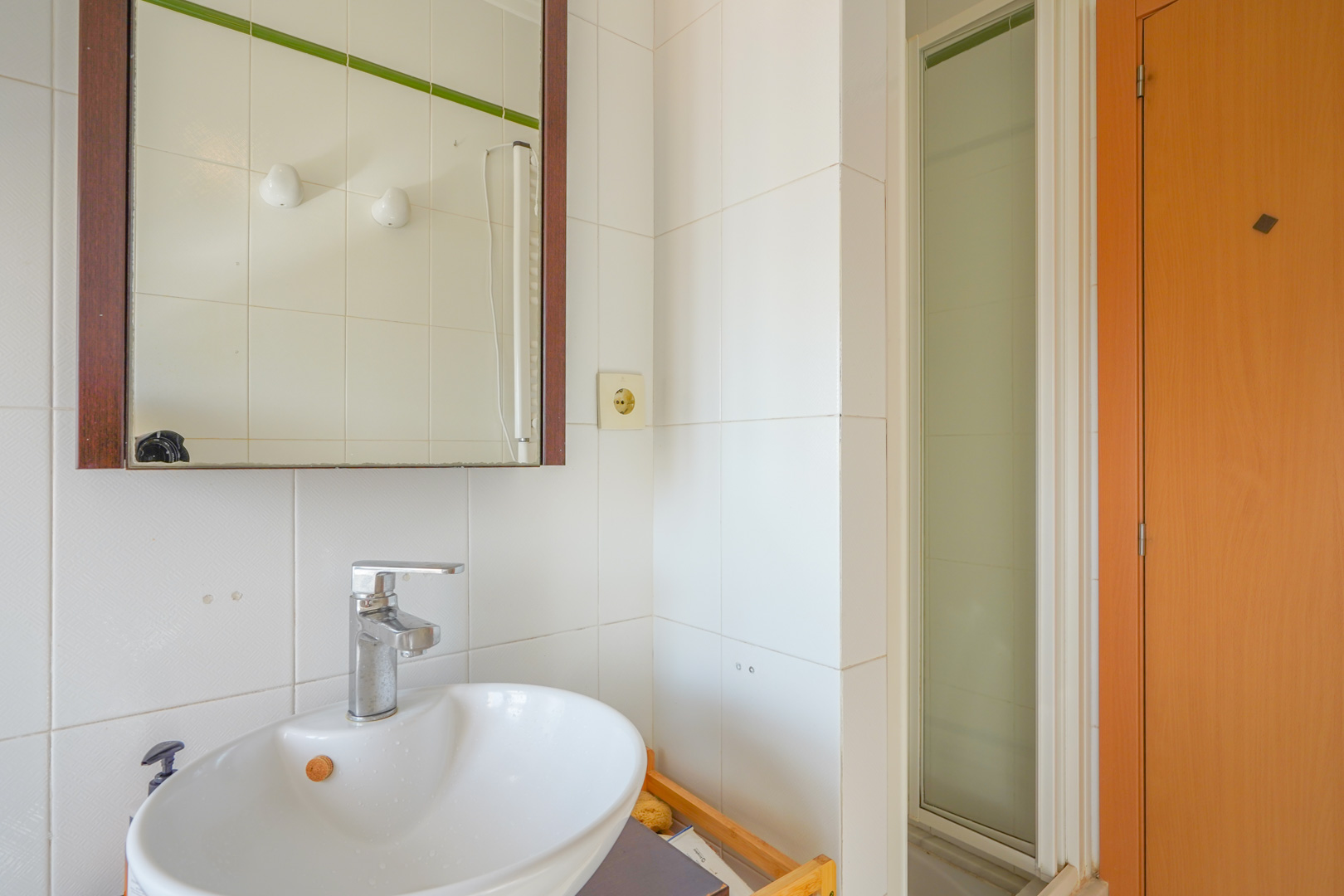 Baño funcional con lavabo de cerámica blanca, grifería cromada y espejo con marco de madera oscura, junto a una ducha con mampara de cristal esmerilado.