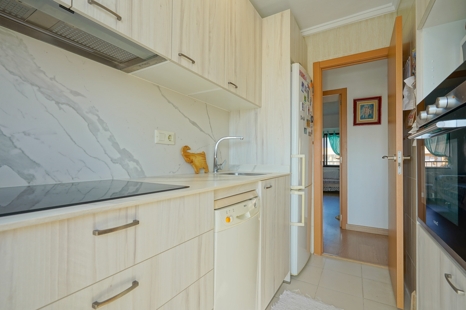 Cocina funcional con armarios de madera clara, frontal de efecto mármol, placa de inducción y electrodomésticos integrados con vista al pasillo.