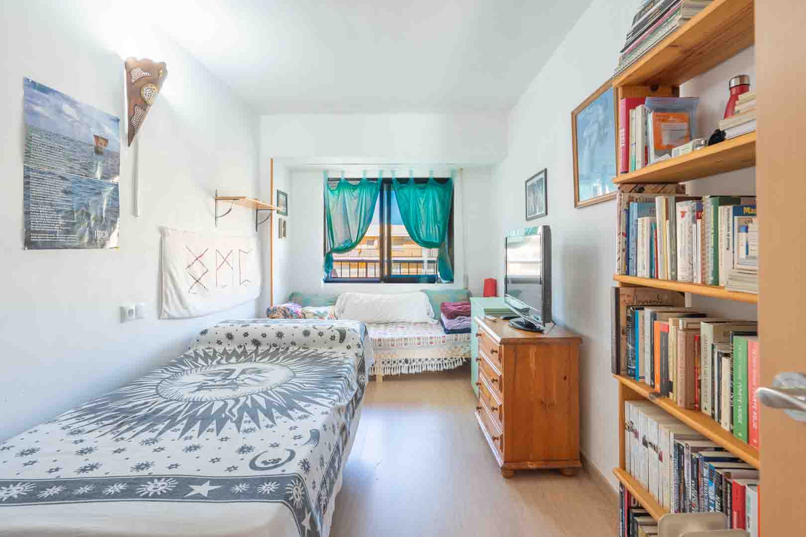 Dormitorio luminoso con cama individual, estantería de madera y cómoda con televisor. La luz natural entra por una ventana vestida con cortinas de color turquesa.