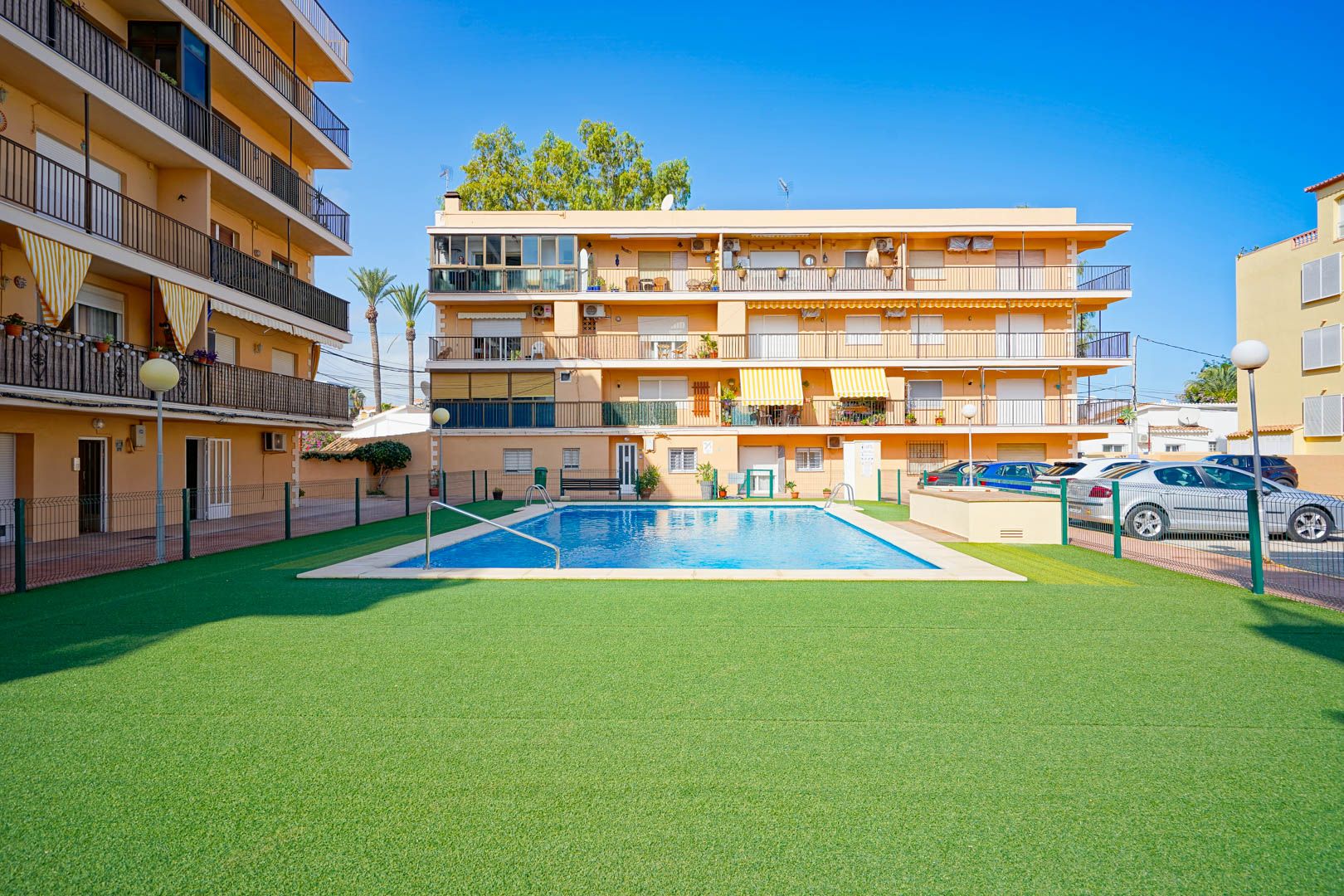Piscina comunitaria rectangular rodeada de una amplia zona de césped artificial y vallado perimetral en un complejo residencial bajo un cielo despejado.