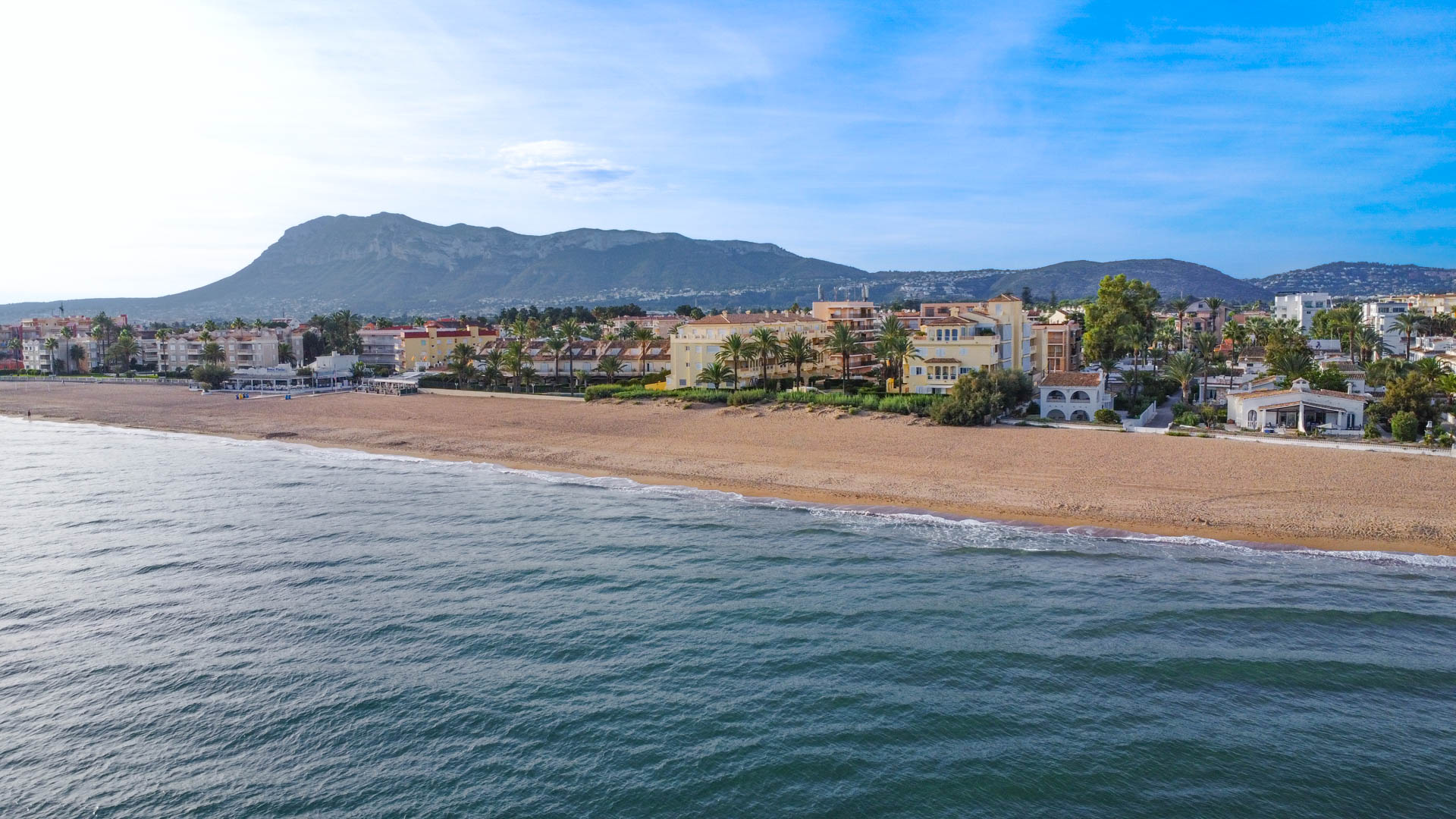 Vista aérea de una playa de arena bordeada por complejos residenciales y palmeras, con una prominente cordillera montañosa al fondo bajo un cielo despejado.