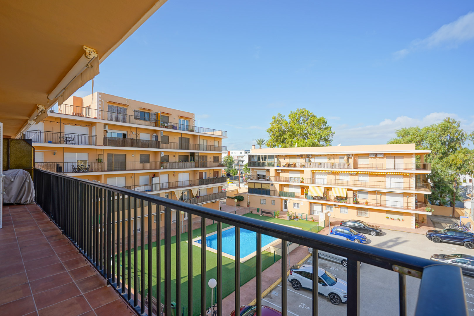 Vista desde un balcón privado con suelo de baldosas y barandilla metálica hacia la piscina comunitaria, zona de césped artificial y aparcamiento del complejo residencial.