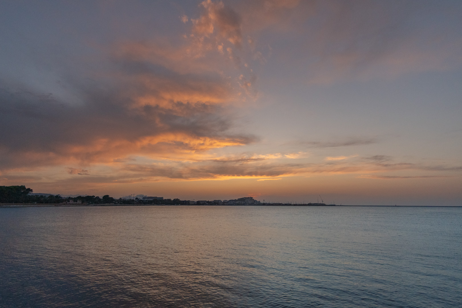 Vista panorámica de la costa mediterránea al atardecer, con nubes en tonos anaranjados y aguas tranquilas que reflejan el cielo sobre la silueta urbana lejana.