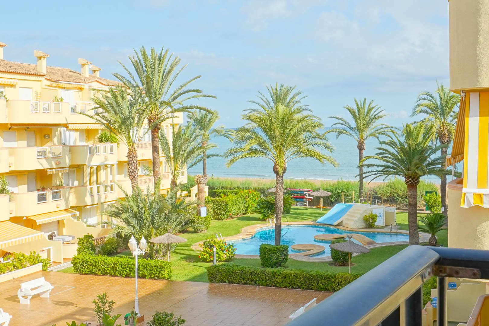 Vistas desde un balcón privado hacia el jardín comunitario con palmeras, piscina con tobogán y acceso directo a la playa del Mediterráneo.