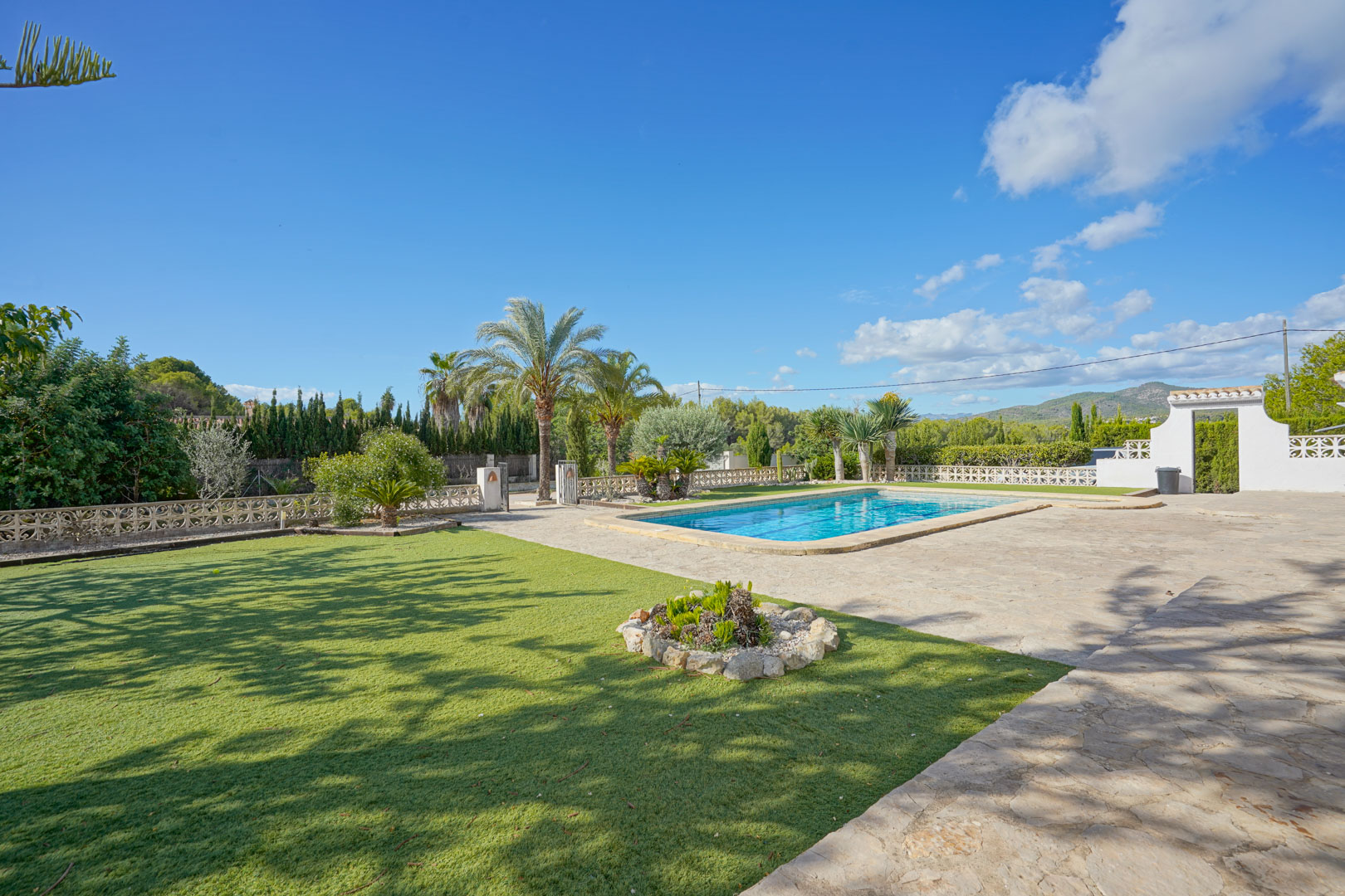 Amplia zona exterior con piscina rectangular, rodeada de una terraza de piedra natural y un jardín cuidado con césped y palmeras bajo un cielo despejado.
