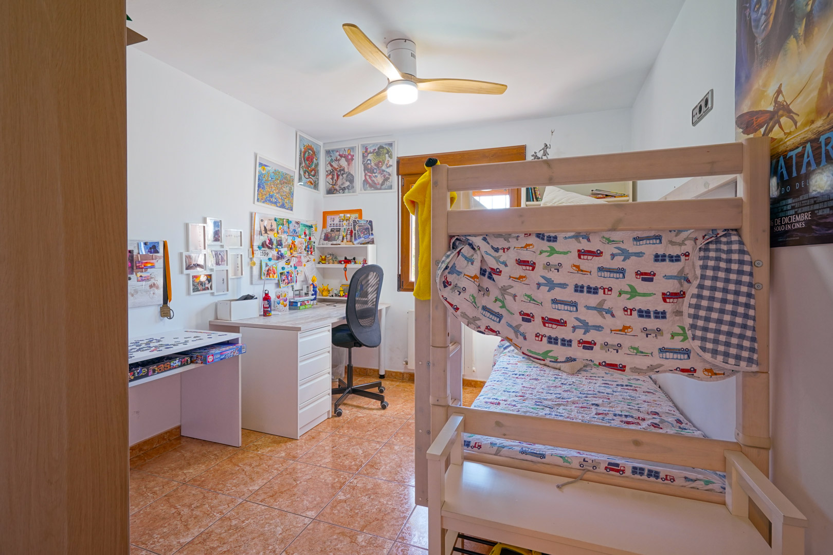 Dormitorio juvenil con literas de madera, zona de estudio con escritorio y ventilador de techo. Incluye suelos de gres y luz natural proveniente de una ventana con marco de madera.