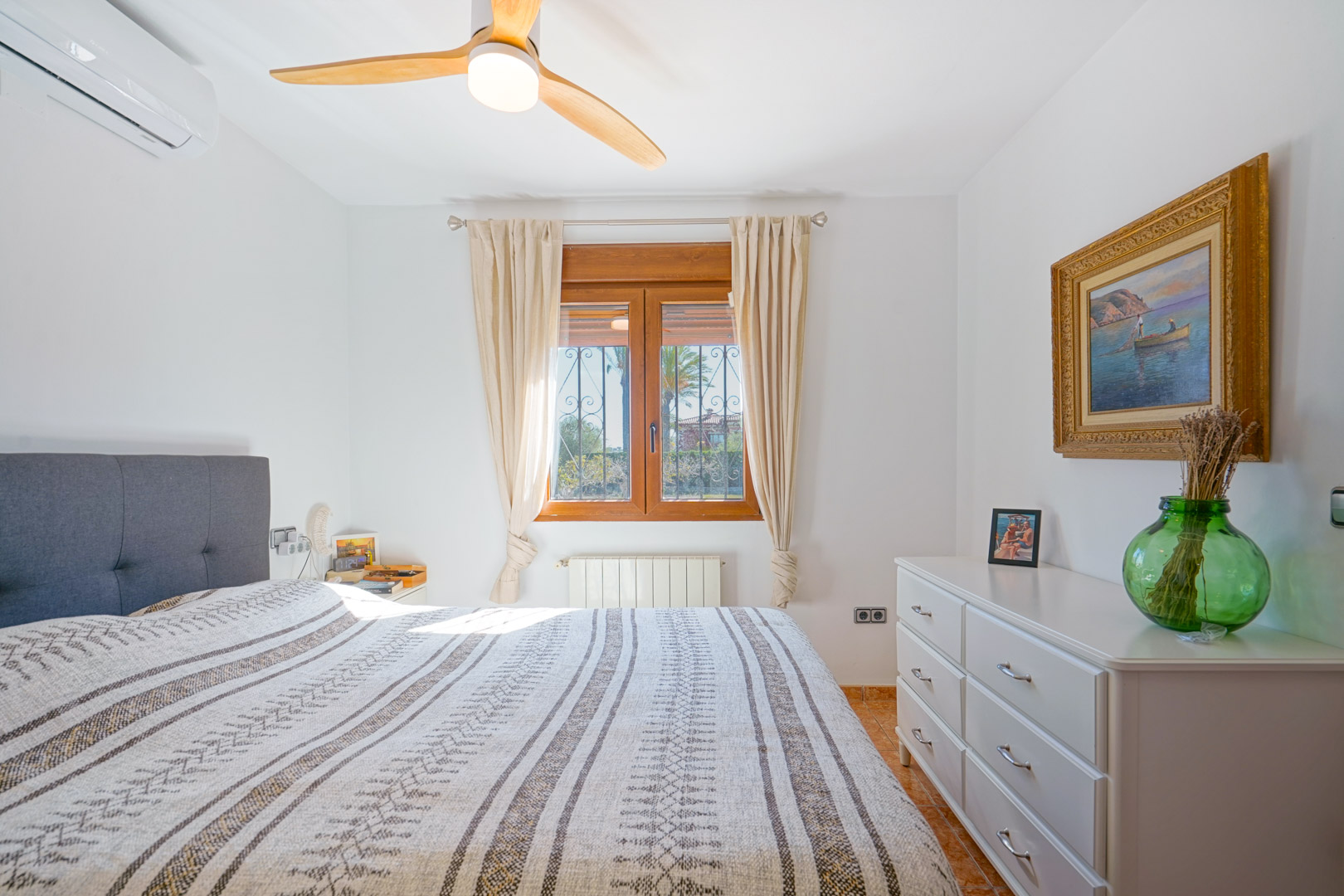Dormitorio luminoso con ventana de madera, ventilador de techo de aspas de madera, aire acondicionado y cómoda blanca. Incluye calefacción por radiador y suelos cerámicos.