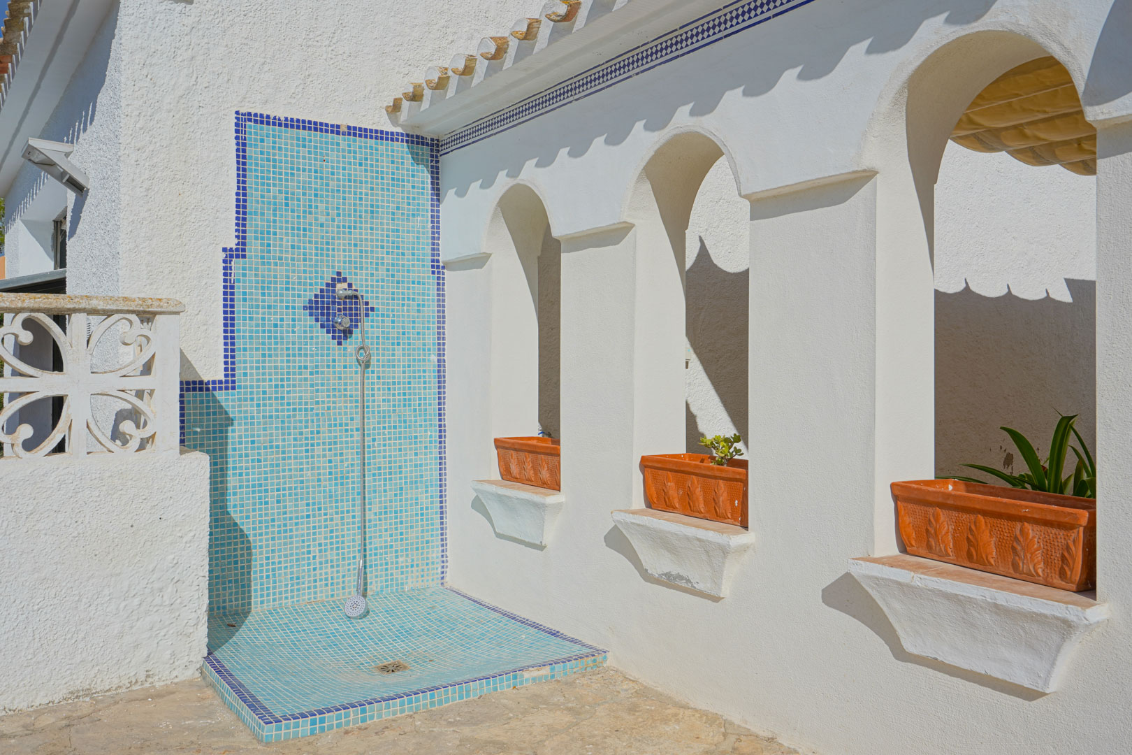 Ducha exterior revestida con mosaico azul junto a una pared de arcos mediterráneos y maceteros de terracota integrados.