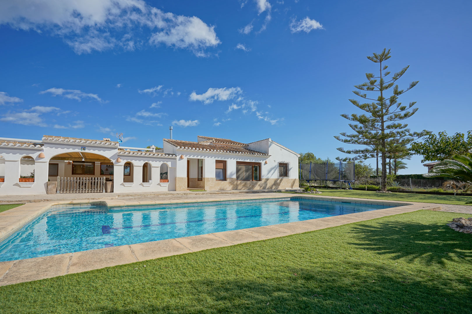 Exterior de la villa con piscina rectangular, rodeada de césped natural y terraza de piedra. Destaca la arquitectura blanca con porches de arcos y tejados de teja cerámica.