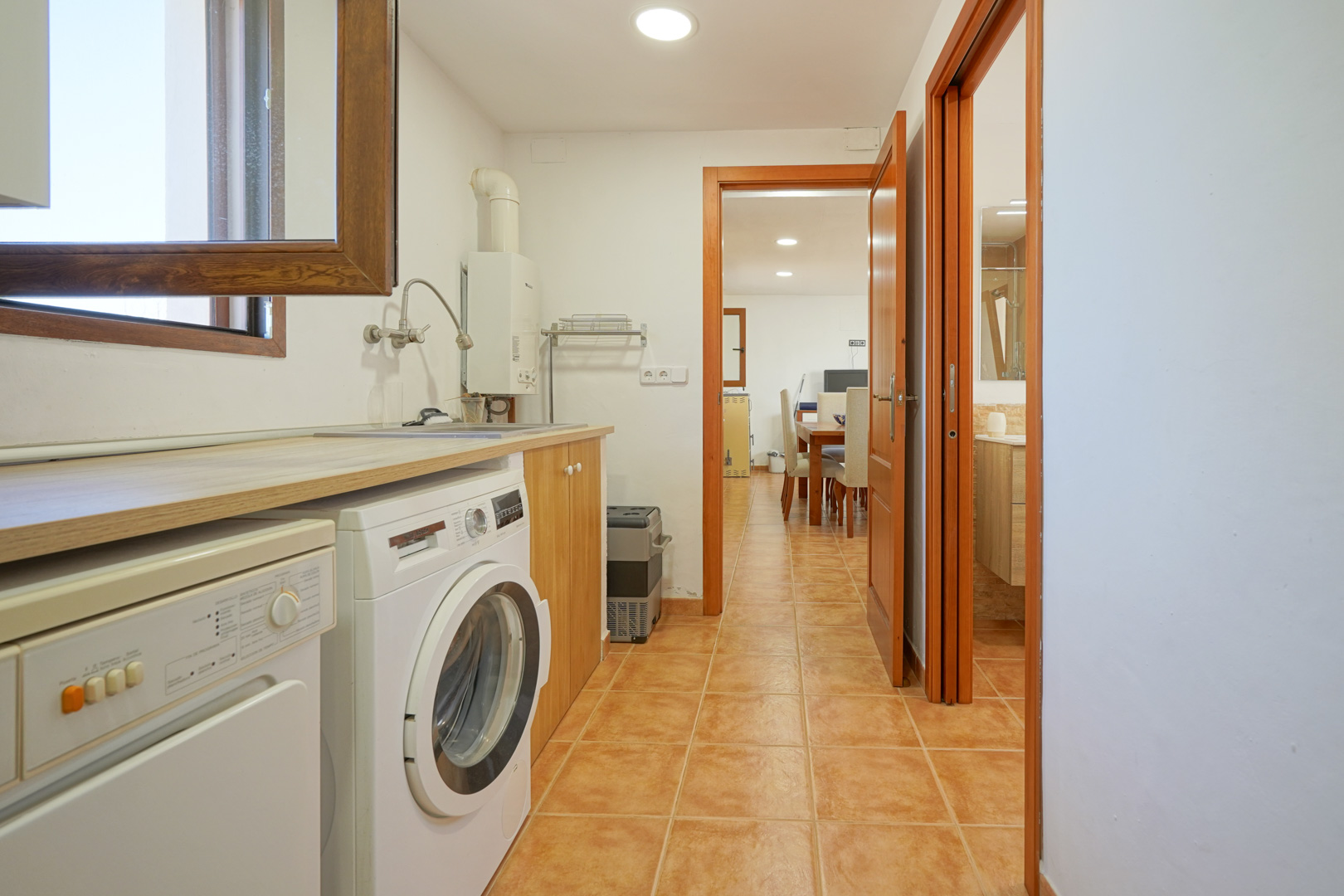 Lavadero funcional con encimera de madera, lavadora y fregadero, conectado directamente con el salón y un baño mediante suelos de gres rústico.