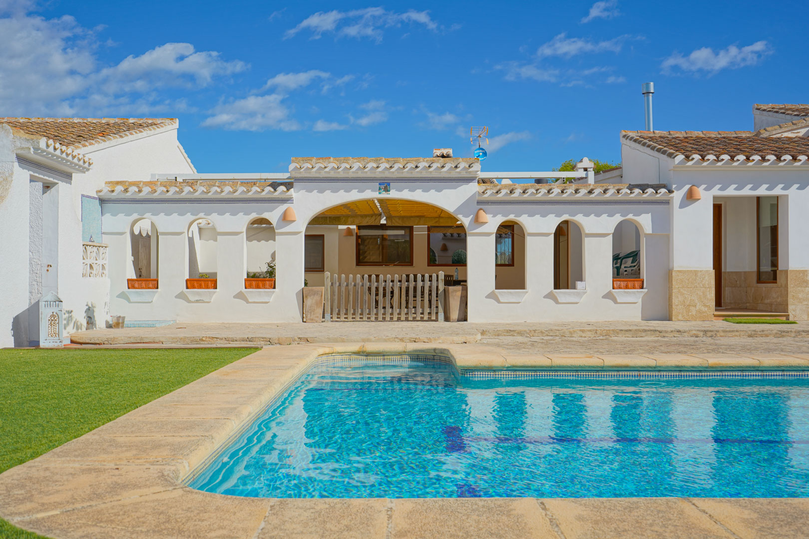 Piscina exterior con borde de piedra natural y terraza soleada frente a una fachada blanca de estilo mediterráneo con arcos tradicionales y tejado de teja.