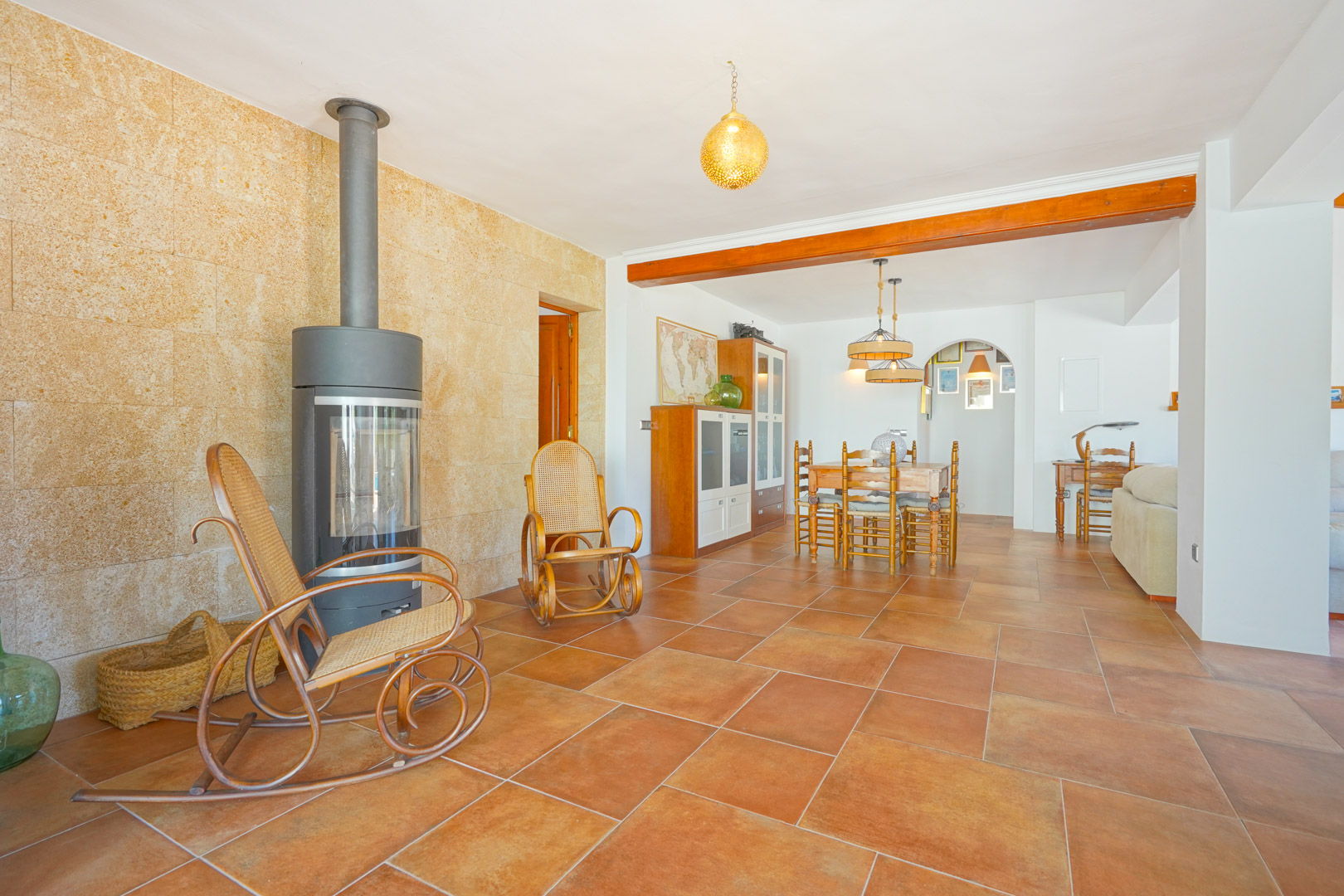 Salón con estufa de leña moderna sobre pared de piedra natural, suelos de terracota y zona de comedor integrada bajo vigas de madera vistas.