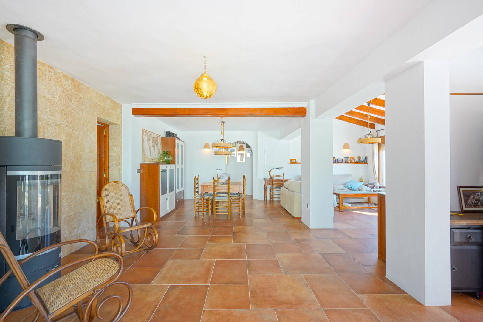 Salón de planta abierta con suelos de terracota, una estufa de leña moderna sobre pared de piedra y vigas de madera vistas que delimitan el comedor.