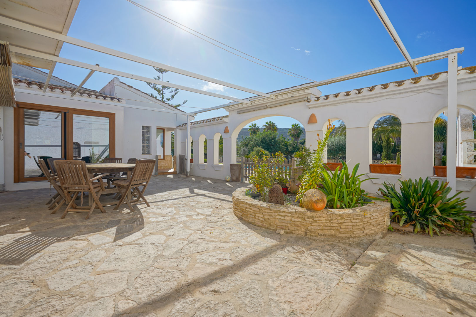 Terraza exterior con suelo de piedra natural, zona de comedor de madera y jardinera circular integrada bajo una estructura de pérgola metálica.