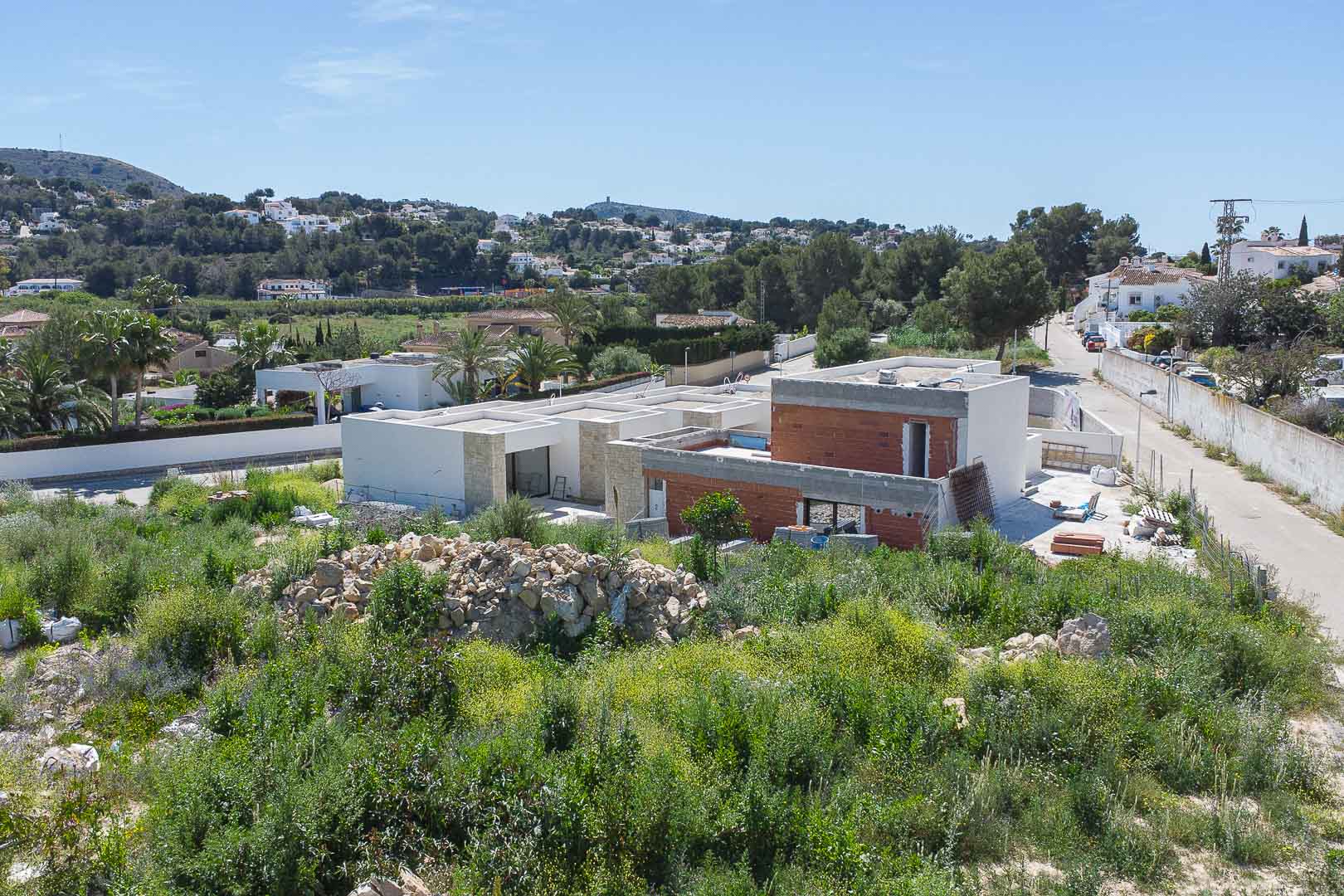Vista exterior de la propiedad en construcción, destacando su arquitectura de líneas rectas, fachadas de piedra natural y muros de ladrillo en un entorno mediterráneo.