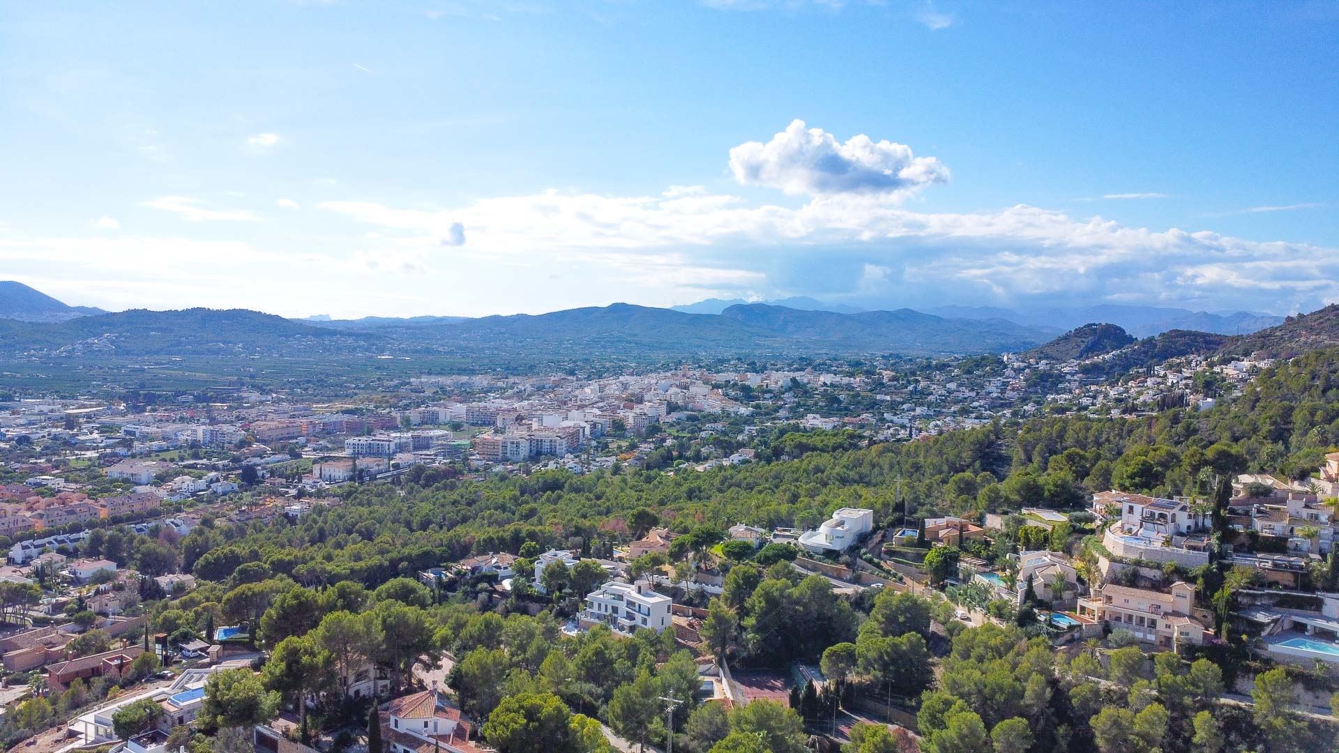 Vista aérea de la urbanización La Corona en Jávea, destacando la integración de villas en la ladera arbolada con vistas despejadas hacia el valle y las montañas.