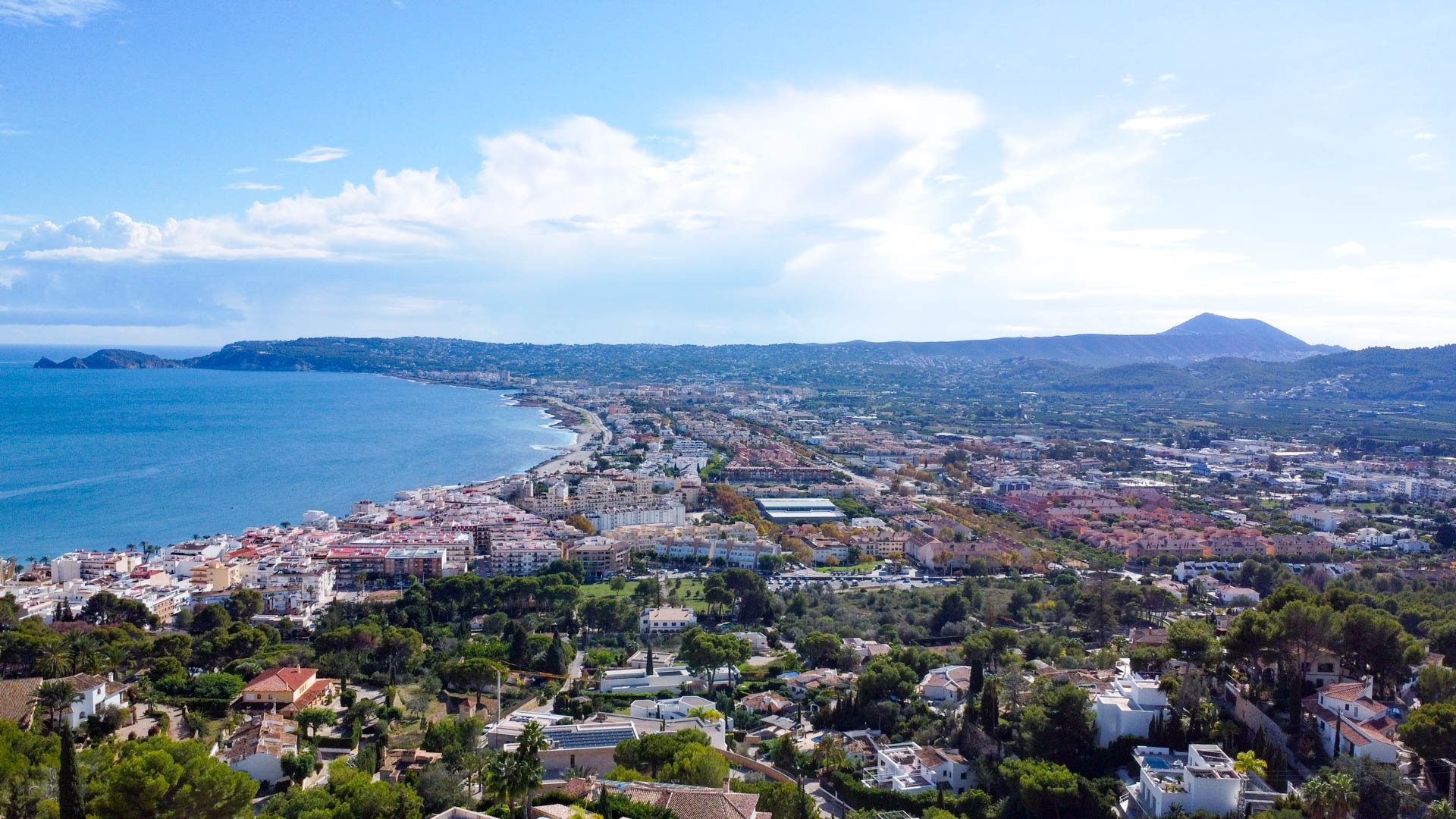 Vista aérea panorámica de la bahía de Jávea y el puerto desde la urbanización La Corona, mostrando la costa mediterránea y el perfil del Montgó al fondo.