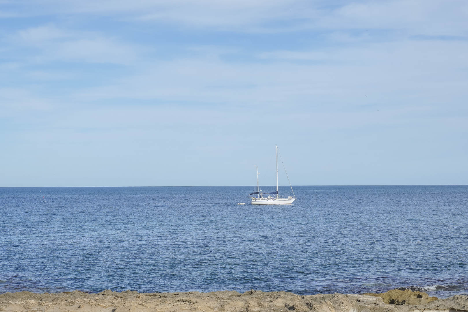 Vistas panorámicas al mar Mediterráneo desde la costa de Jávea, con un velero navegando frente a la costa rocosa bajo un cielo despejado.